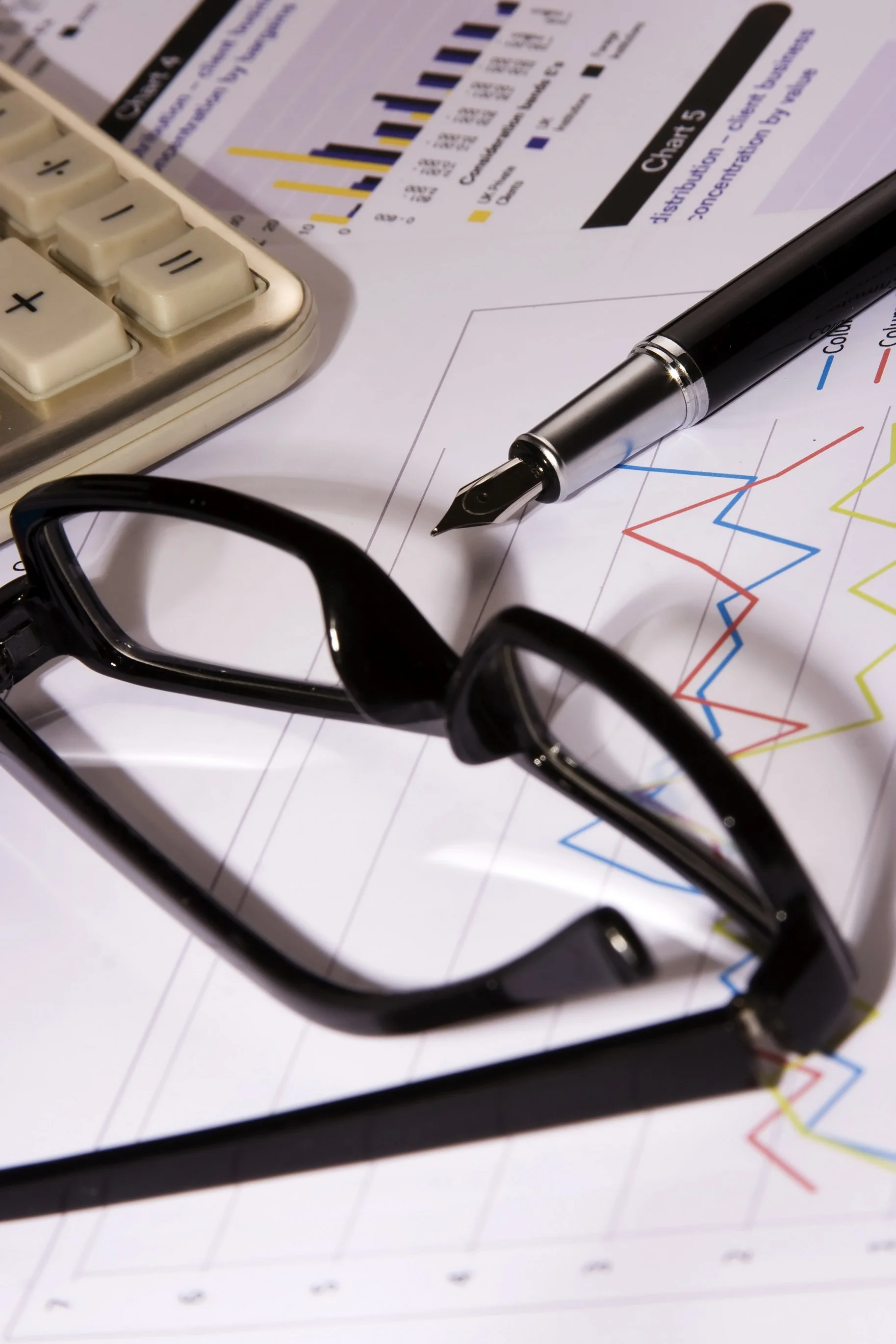 Close-up of a business workspace with a calculator, a fountain pen, glasses, and printed financial graphs and charts.