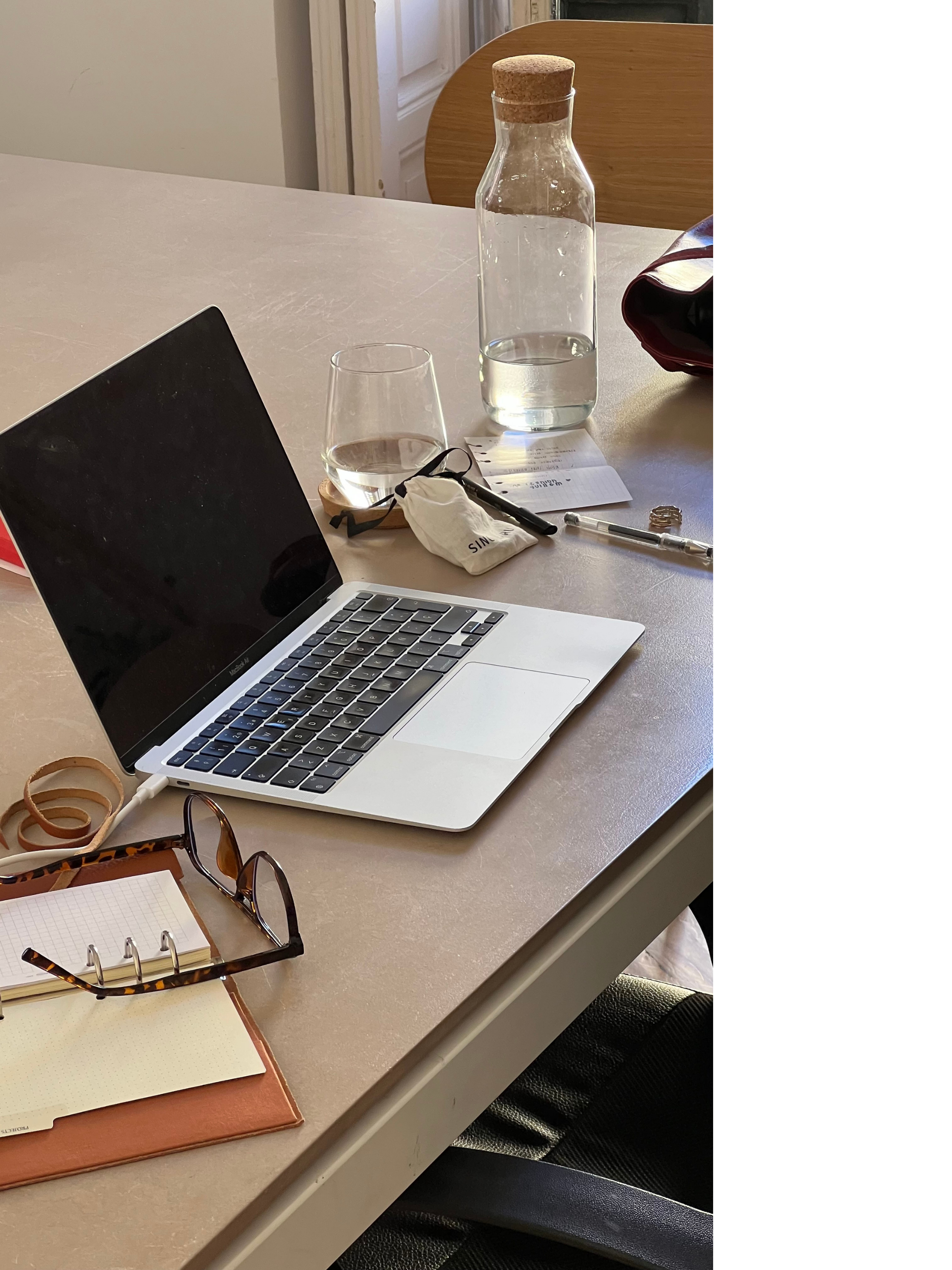 A laptop, glasses, a water bottle, a glass of water, a pen, a notebook, and a pair of glasses on a beige table, with a wooden chair in the background.