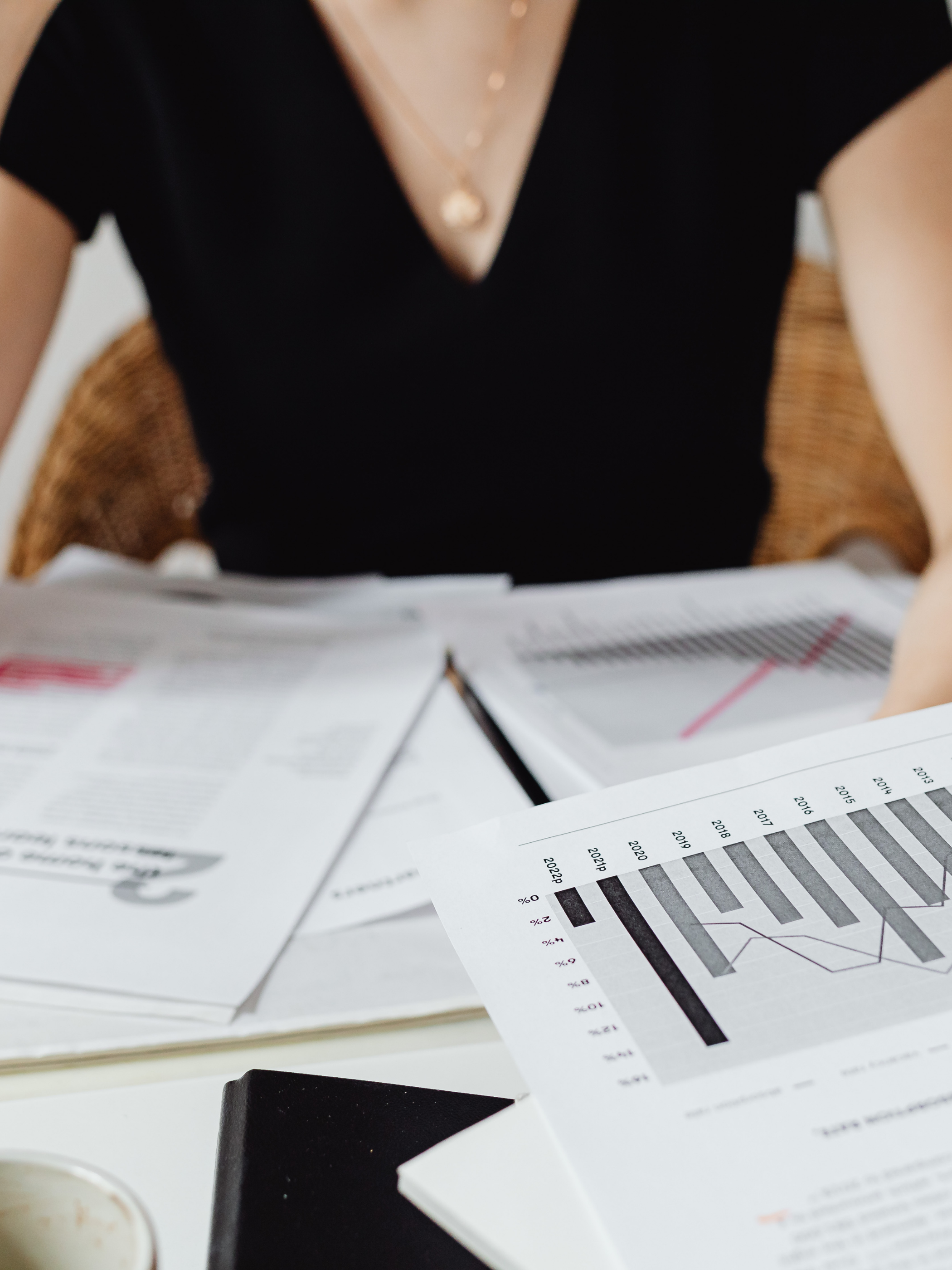Person sitting at a desk with financial documents and charts, wearing a black top and a gold necklace.