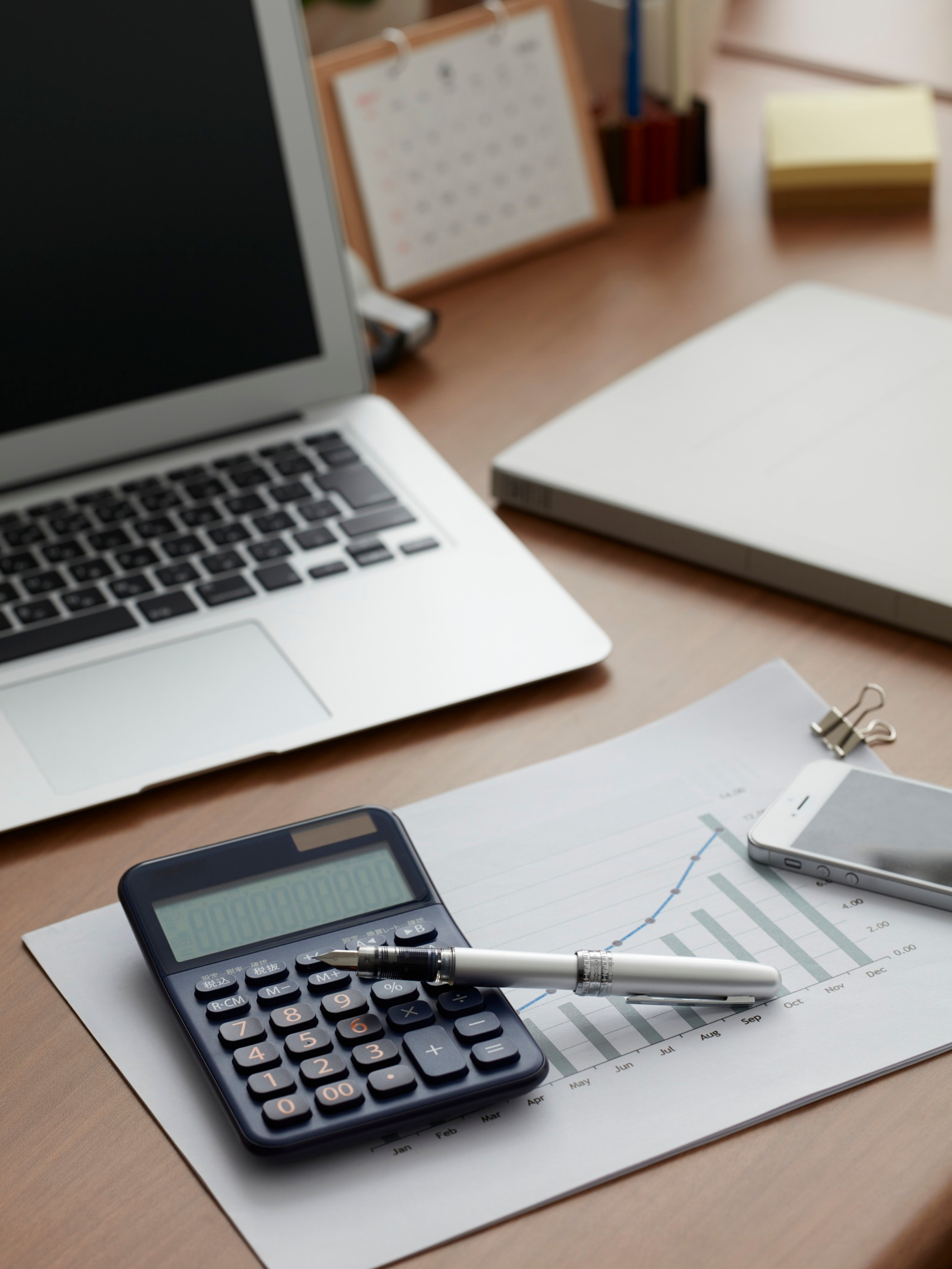 Desk with open laptop, calculator, pen, paper with financial chart, smartphone, and office supplies.