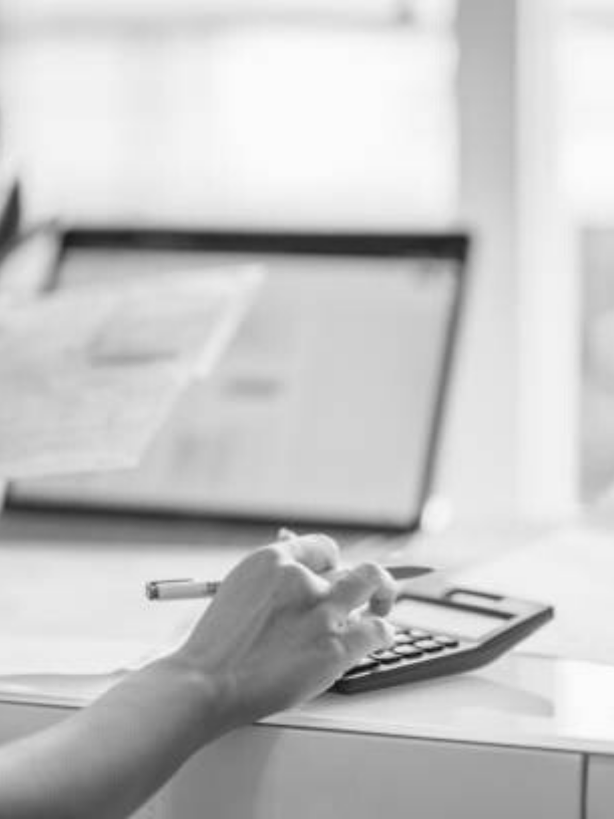 A person's hand holding a pen pressing keys on a calculator on a desk, with a computer screen in the background.