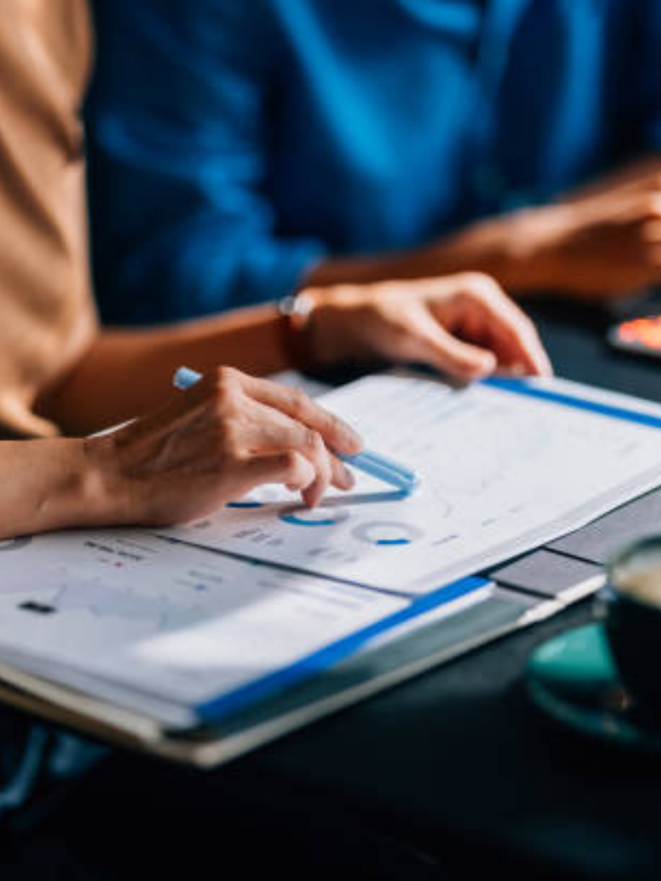 People sitting at a table, working on printed documents or charts, with one person writing or pointing to the paper.
