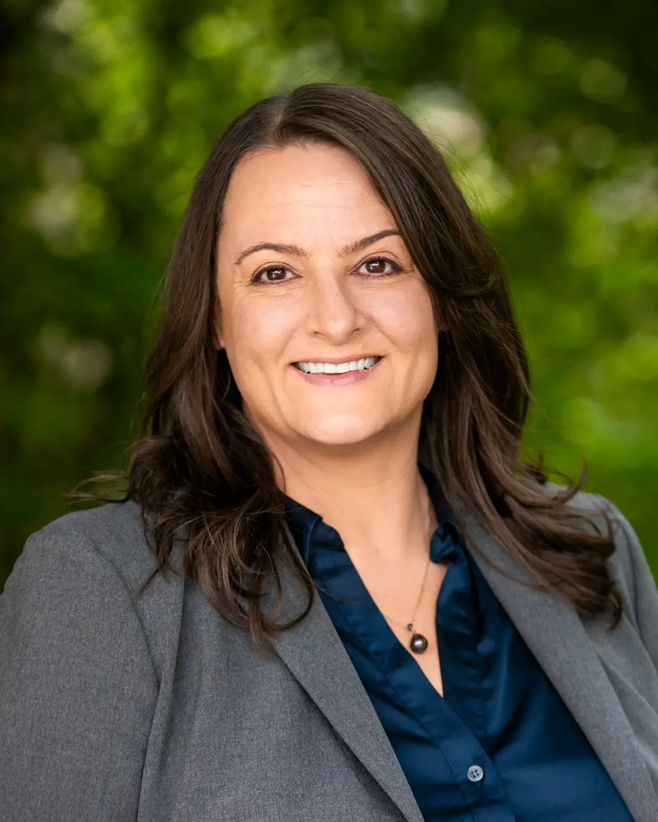 Professional woman with brown hair in a gray blazer and navy blouse smiling outdoors with green blurred background.