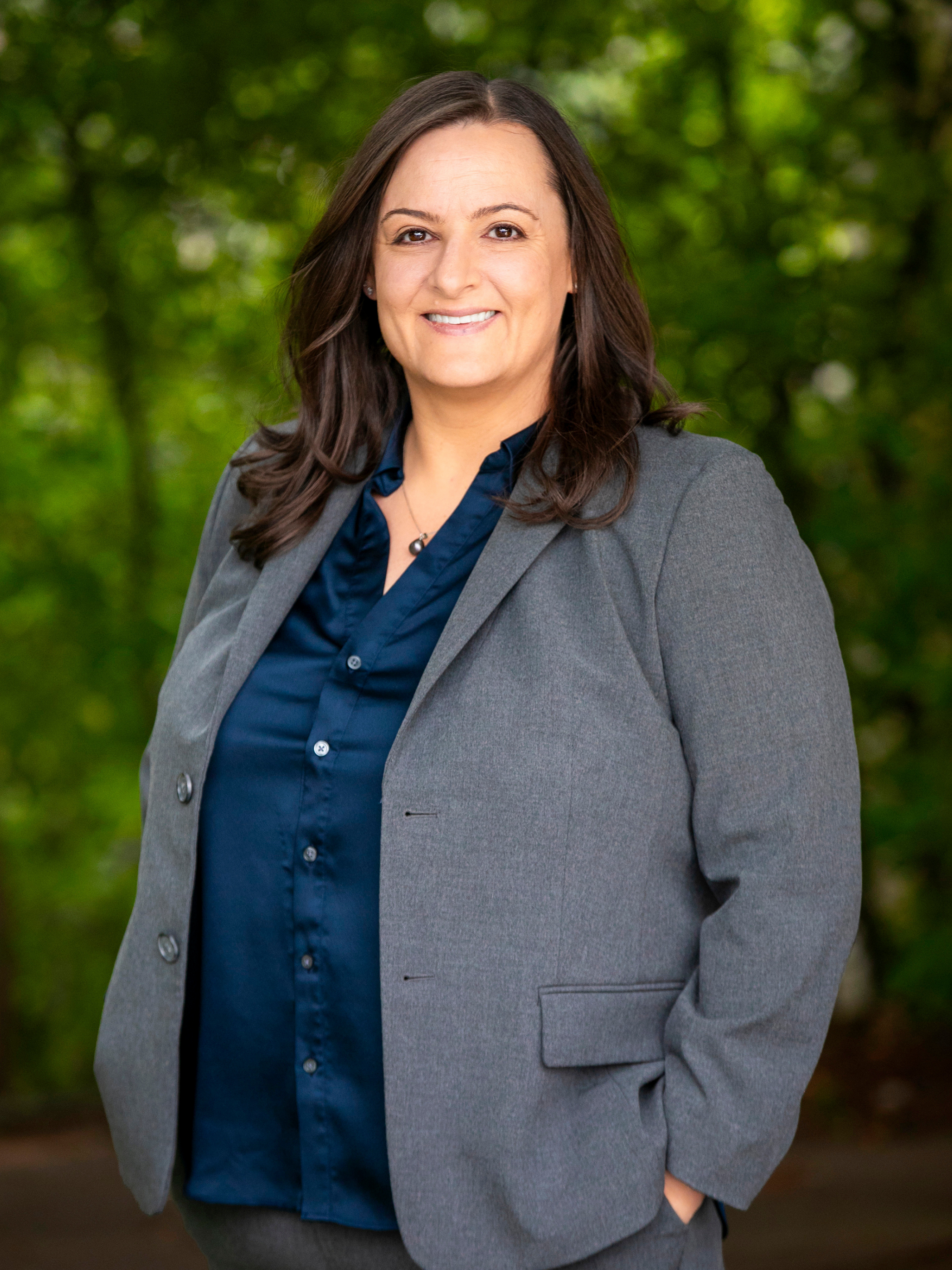 A professional woman with dark brown hair, wearing a gray blazer over a navy blue blouse, smiling outdoors with green foliage in the background.