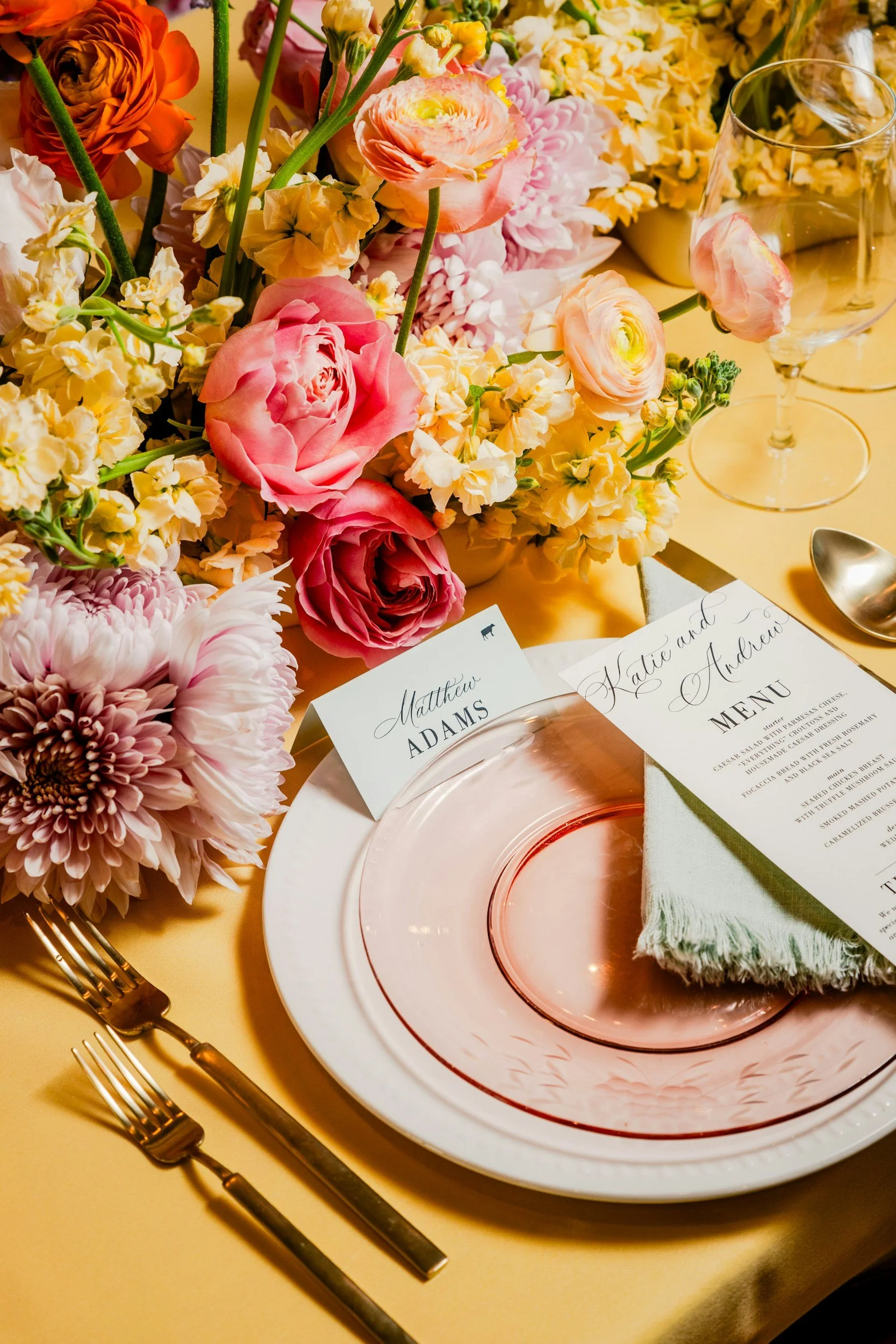 A table setting with a floral centerpiece of pink, peach, and yellow flowers, gold-colored cutlery, a white dinner plate with a pink glass plate on top, a menu, a place card with the name Matthew Adams, a wine glass, and a spoon, all on a yellow tablecloth.