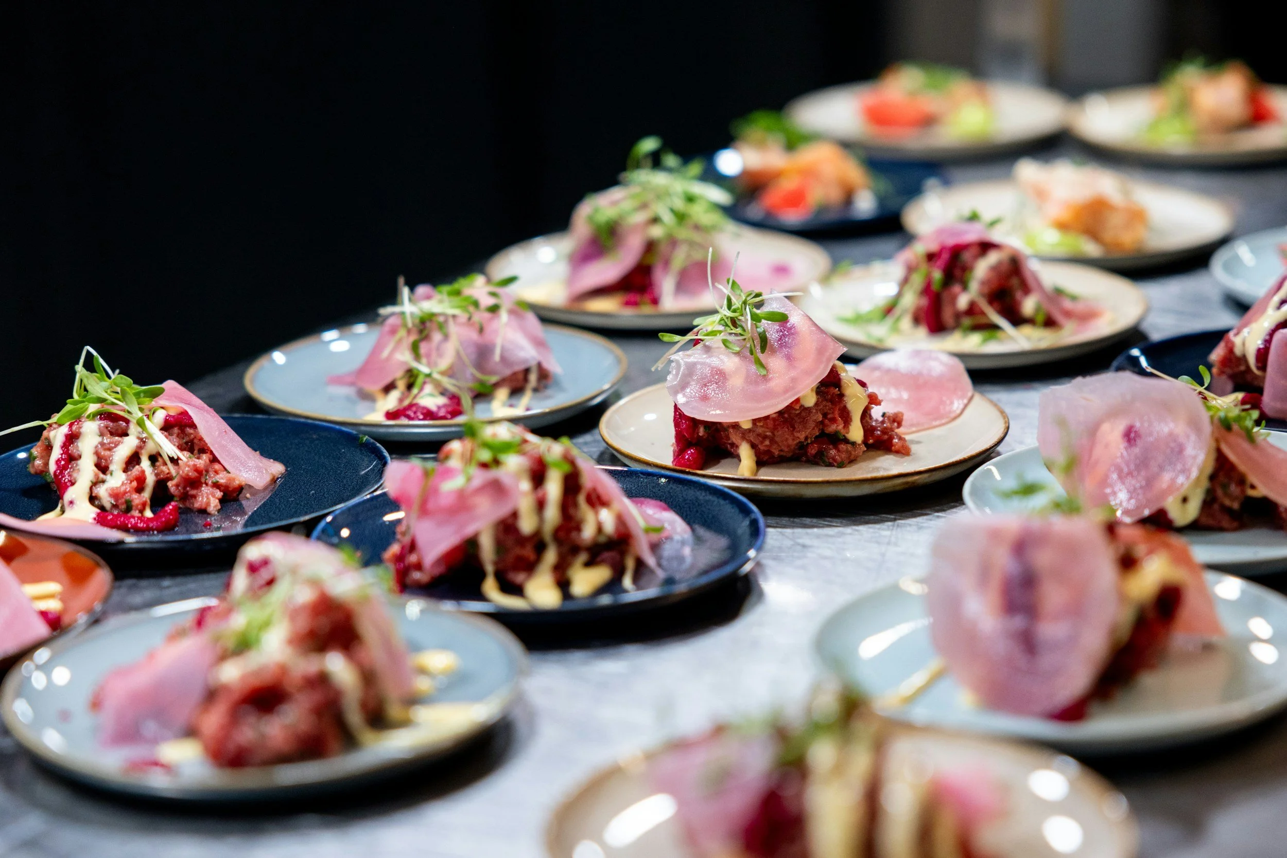 Multiple plates of assorted appetizers with thin slices of pink cured meat, microgreens, and creamy drizzle.