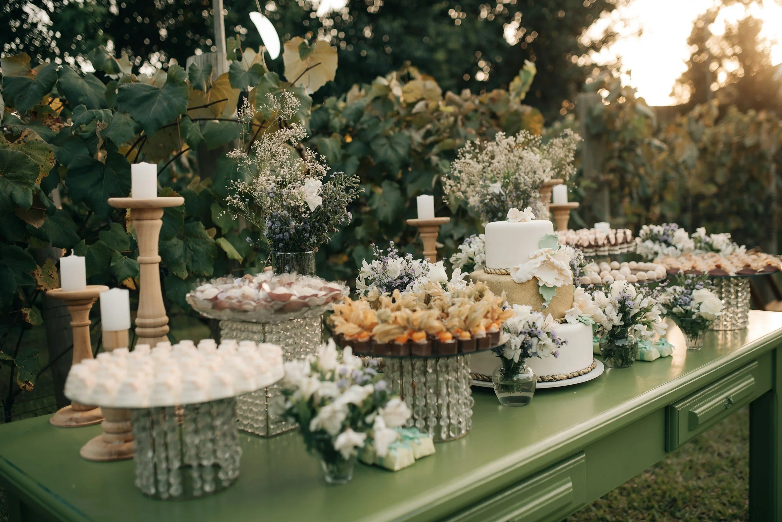 A dessert table decorated with flowers and a wedding cake outdoors during sunset.
