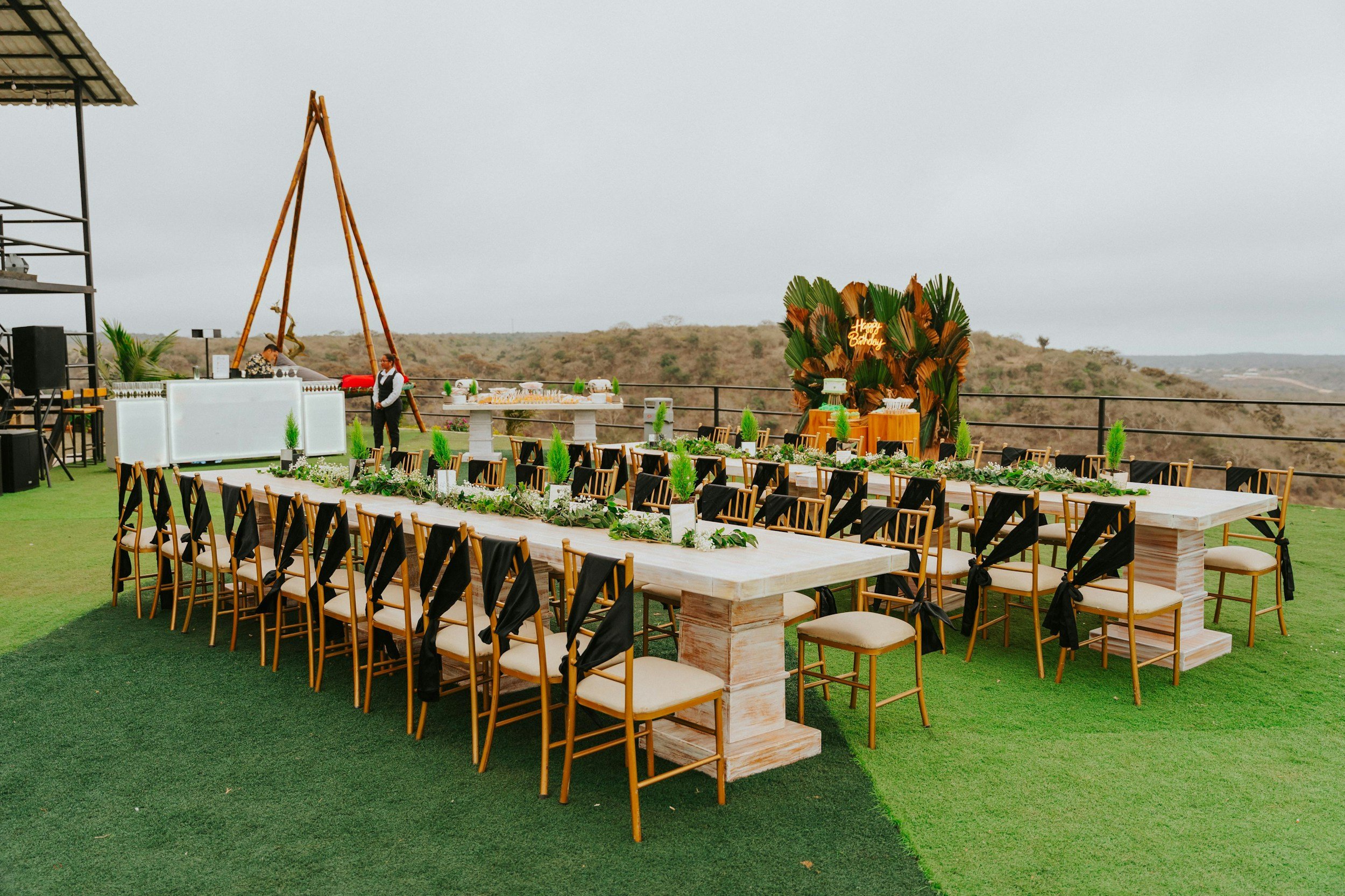 Outdoor event setup with long dining table and chairs, green plant decorations, and a backdrop with a "Happy Birthday" sign amid a scenic landscape on a cloudy day.