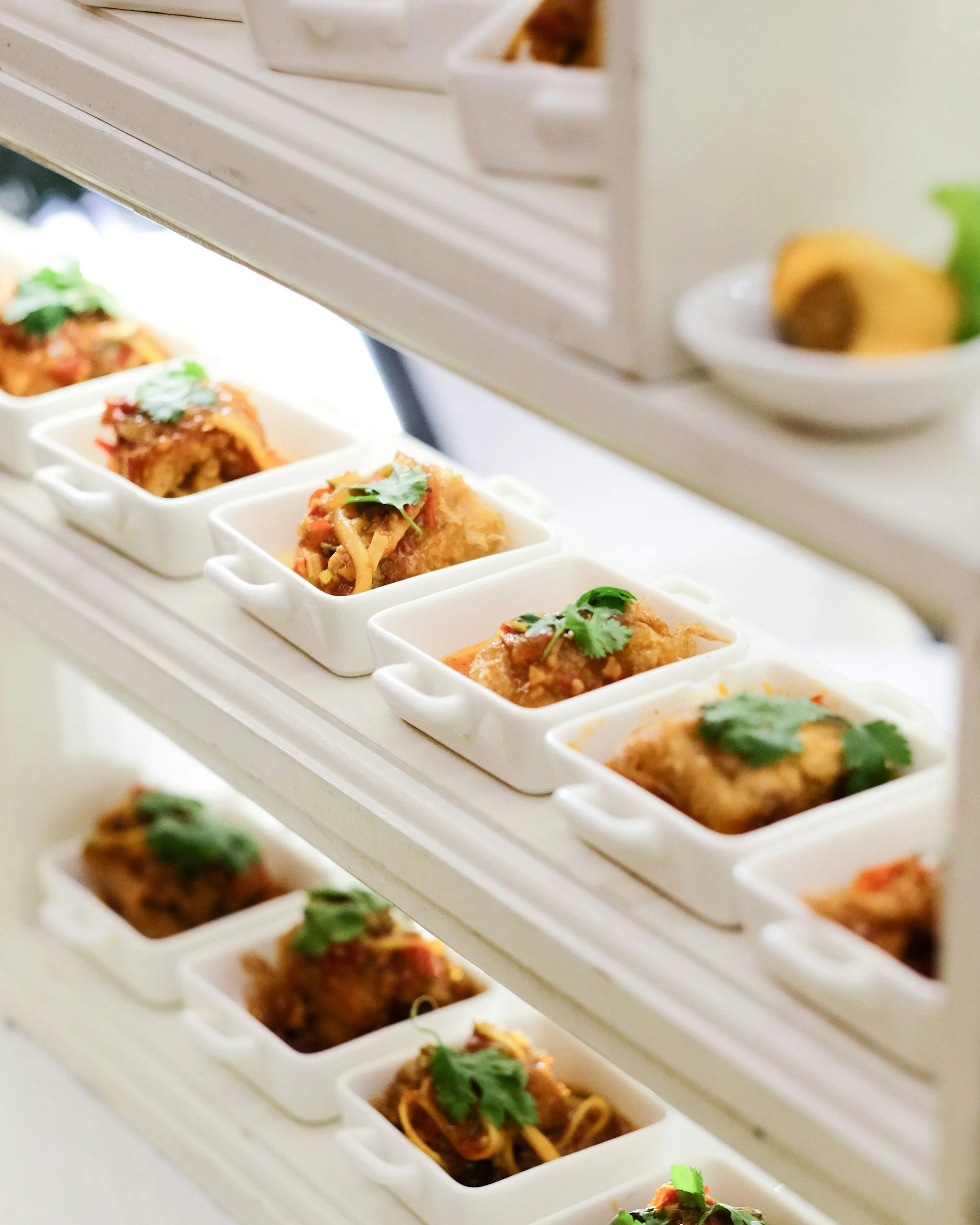 Multiple servings of Asian-style appetizer or entrée, garnished with cilantro, stored in white rectangular dishes on a multi-tiered serving stand.