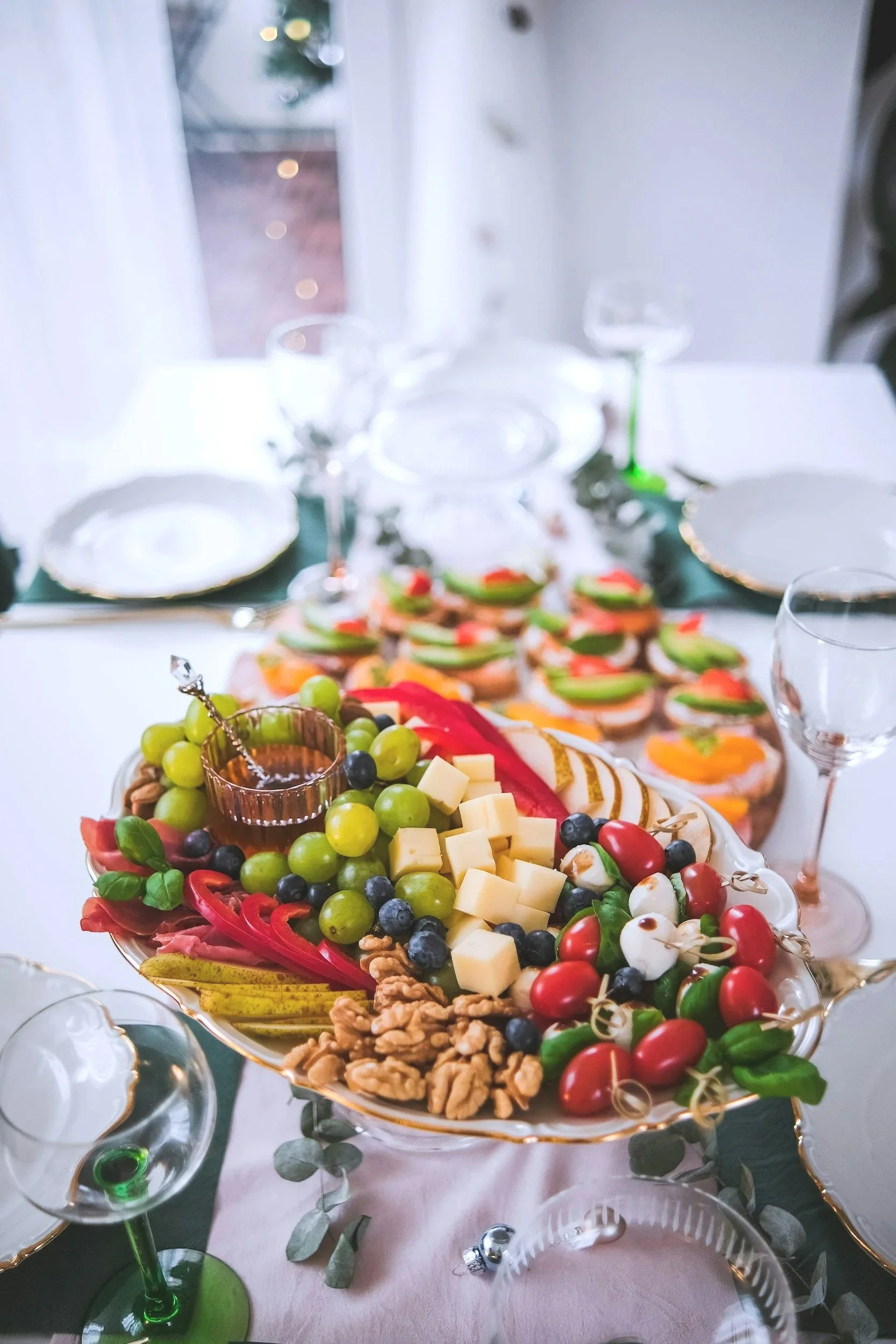 Elegant table setup with a large tray of assorted cheeses, grapes, nuts, and colorful garnished appetizers, surrounded by empty plates and glasses, with a holiday wreath visible through a window in the background.