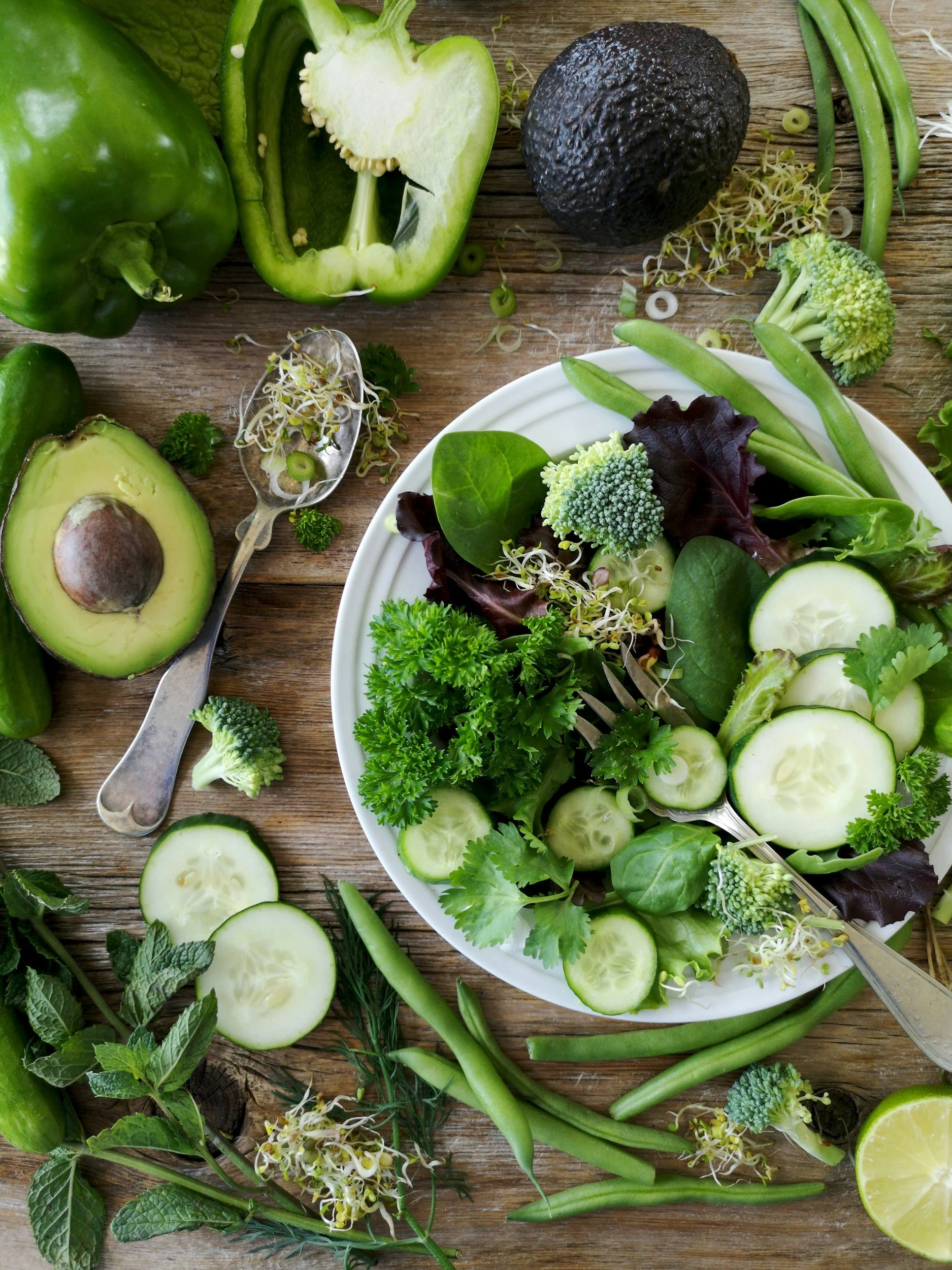 Een overzicht van groene groenten en fruit, waaronder een rode paprika, avocado, komkommer, broccoli, groene bonen, munt en andere bladgroenten, op een houten tafel.