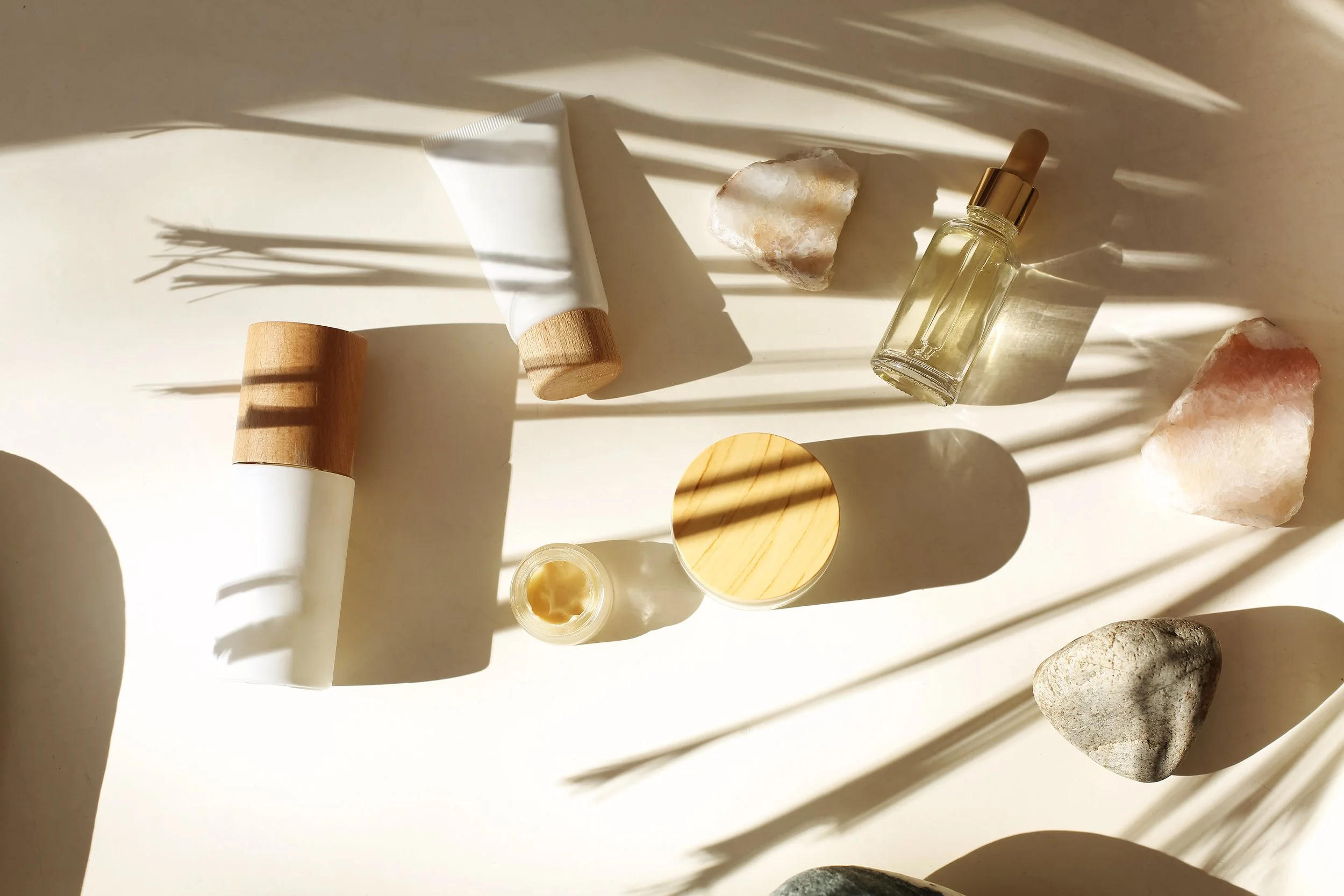 Skincareproducten en stenen op een lichte achtergrond met schaduw van palmbladeren.