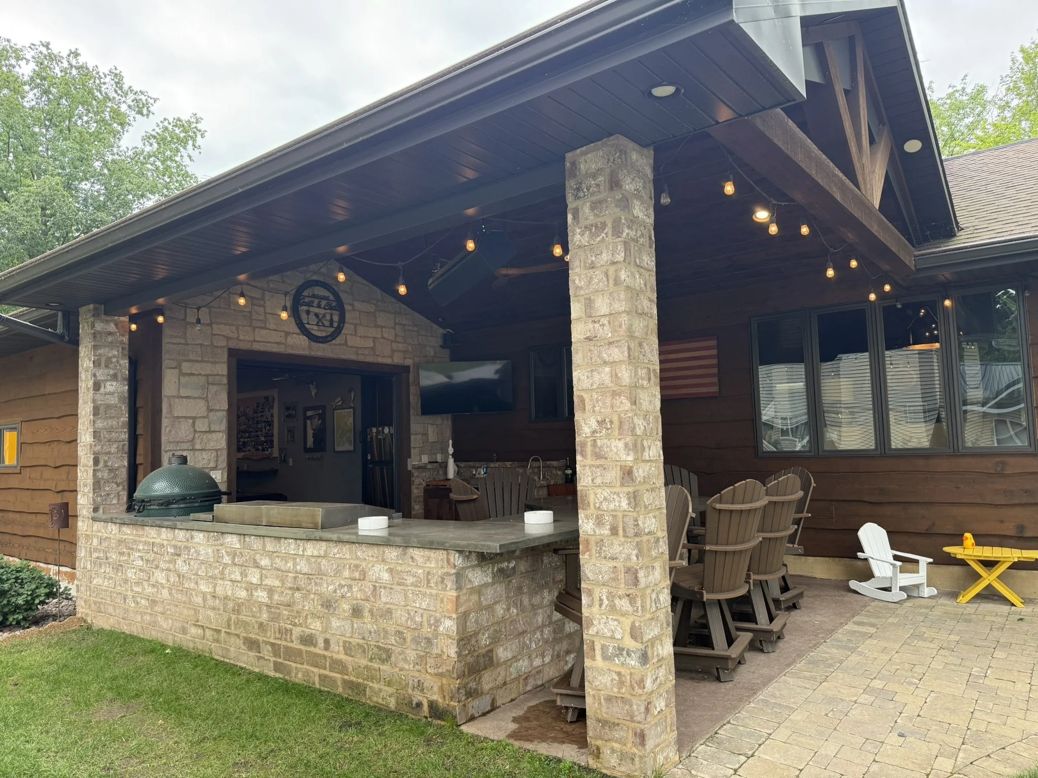 Full build modern/rustic outdoor kitchen, with timberwork, brick bar/peninsula, stained concrete counters, built in big green egg, steel cooktop, char broiler and burners, built-in stainless-steel cabinets, stone clad accent wall, stamped concrete fl
