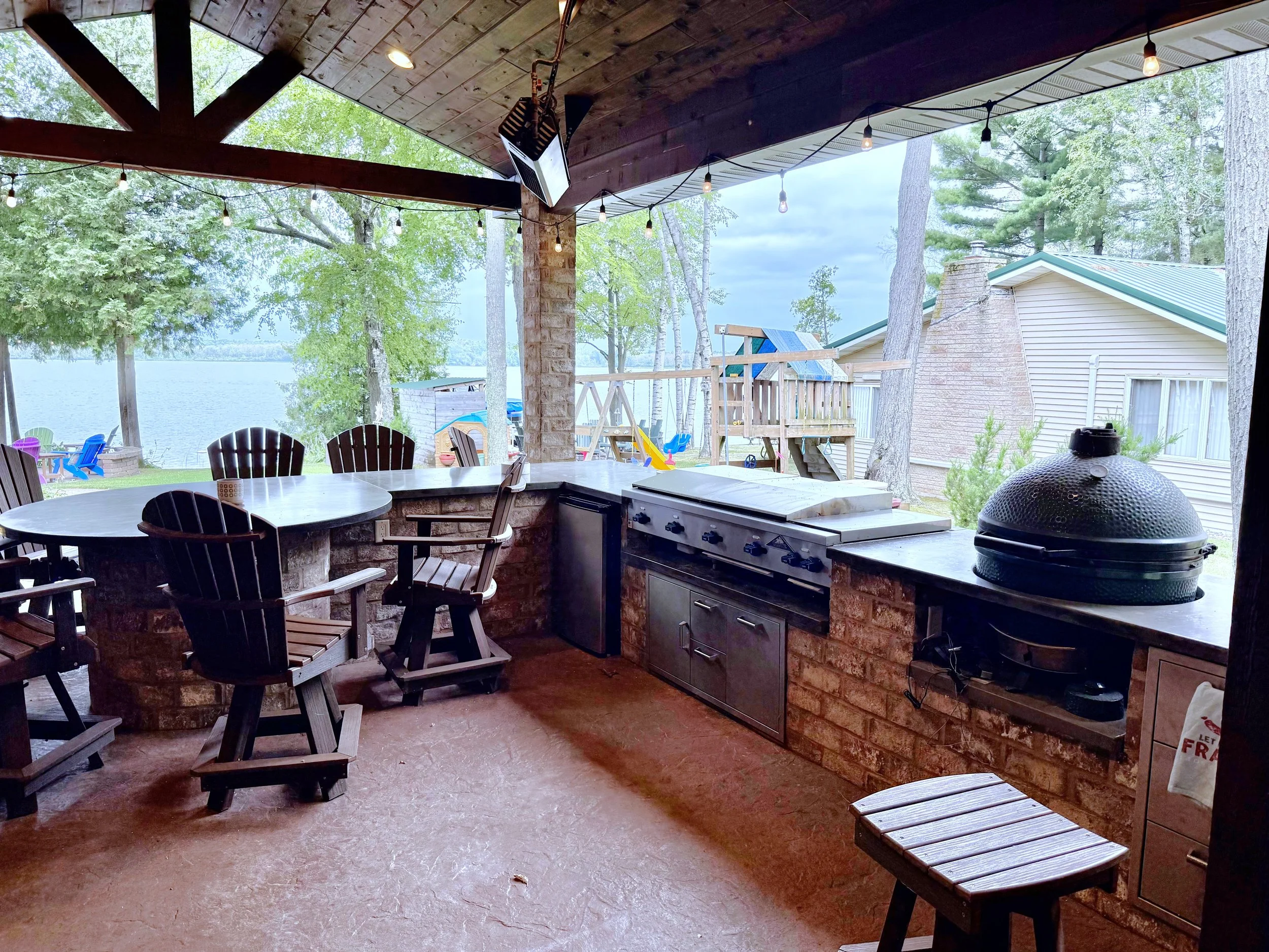 Full build modern/rustic outdoor kitchen, with timberwork, brick bar/peninsula, stained concrete counters, built in big green egg, steel cooktop, char broiler and burners, built-in stainless-steel cabinets, stone clad accent wall, stamped concrete fl