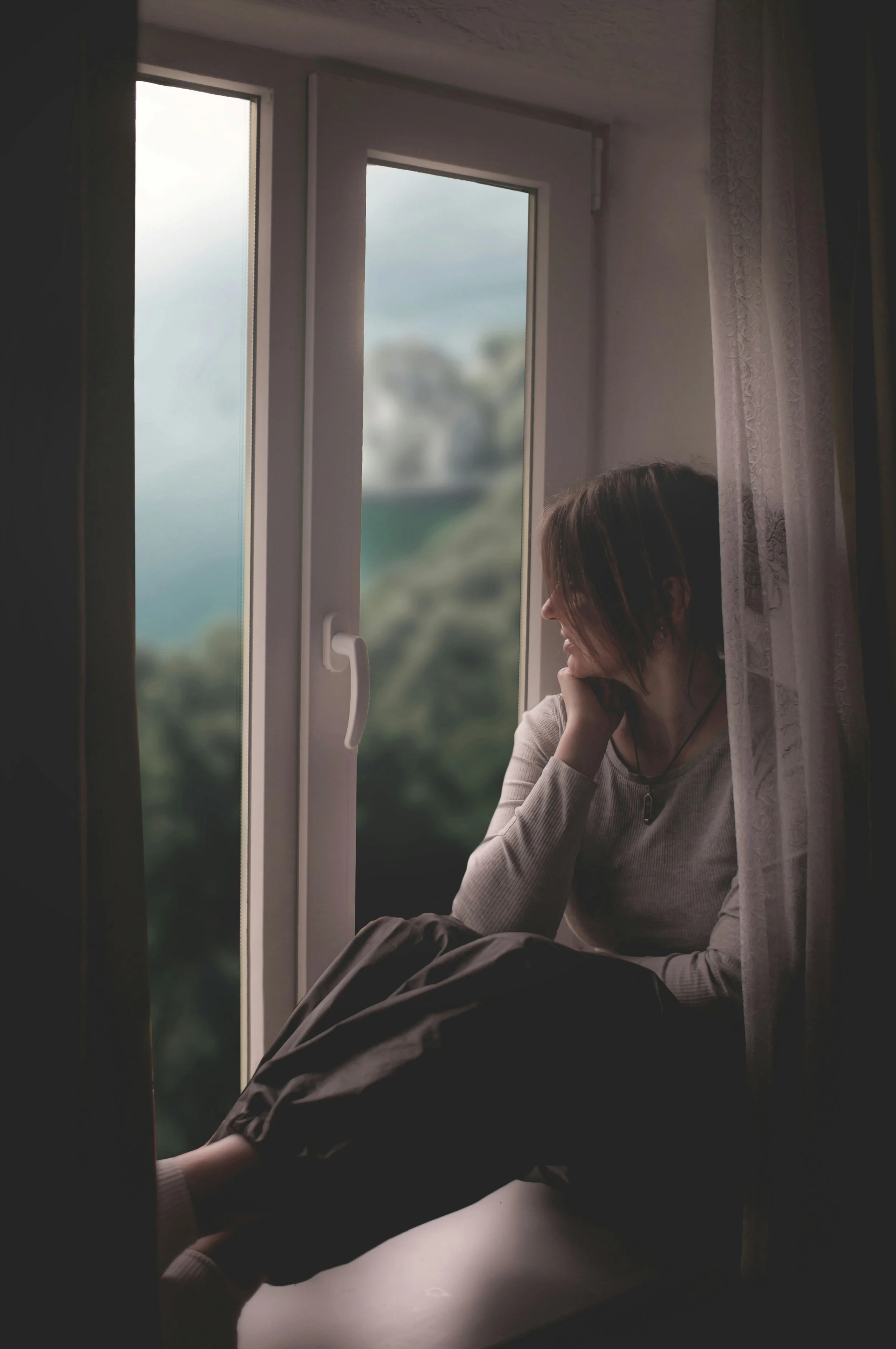 A woman sitting on a windowsill, gazing outside with a peaceful expression, during daytime.