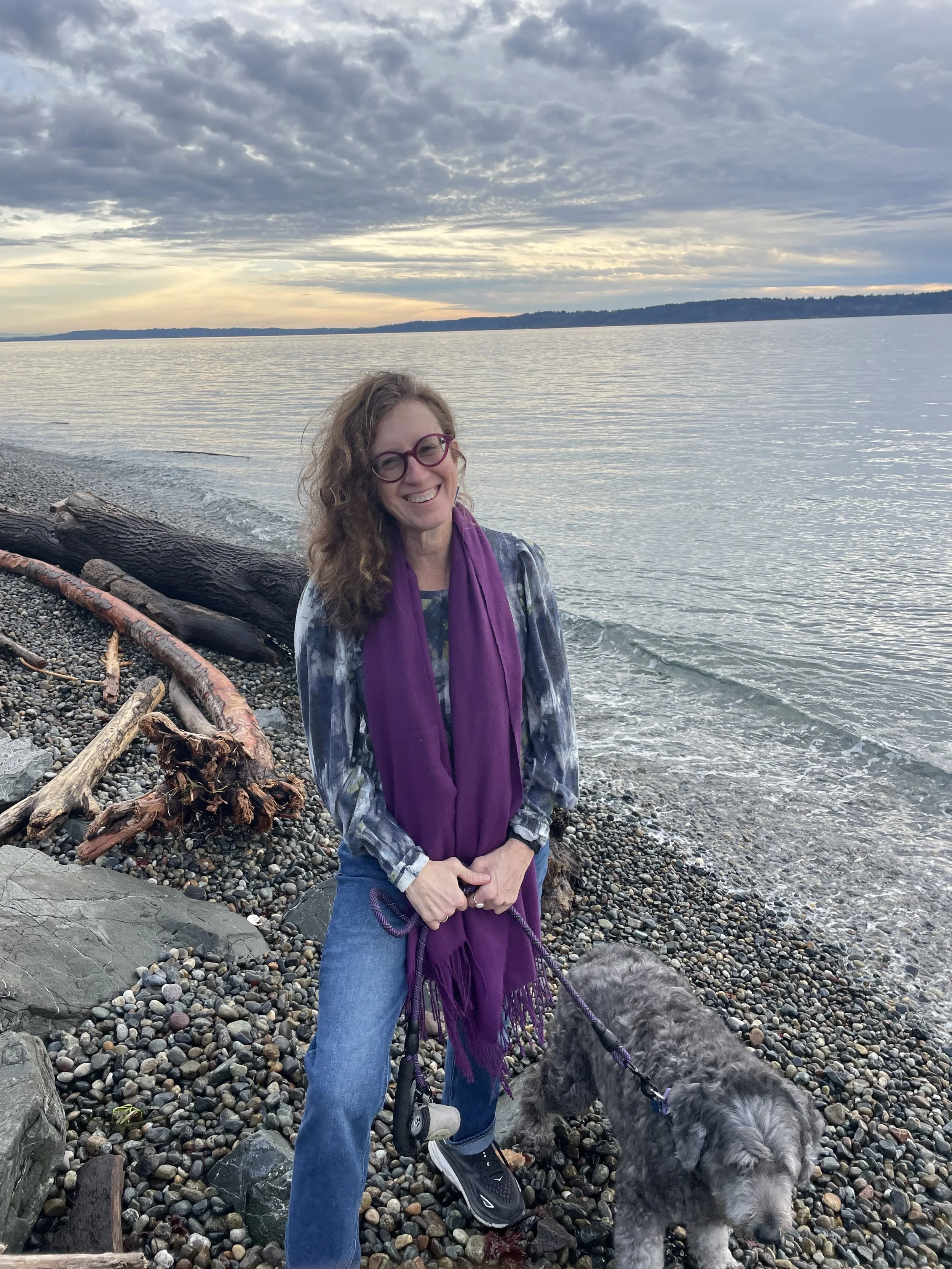 A smiling woman with curly hair and glasses standing on a pebbled beach holding a leash on a curly-haired dog with a lake and cloudy sky in the background.
