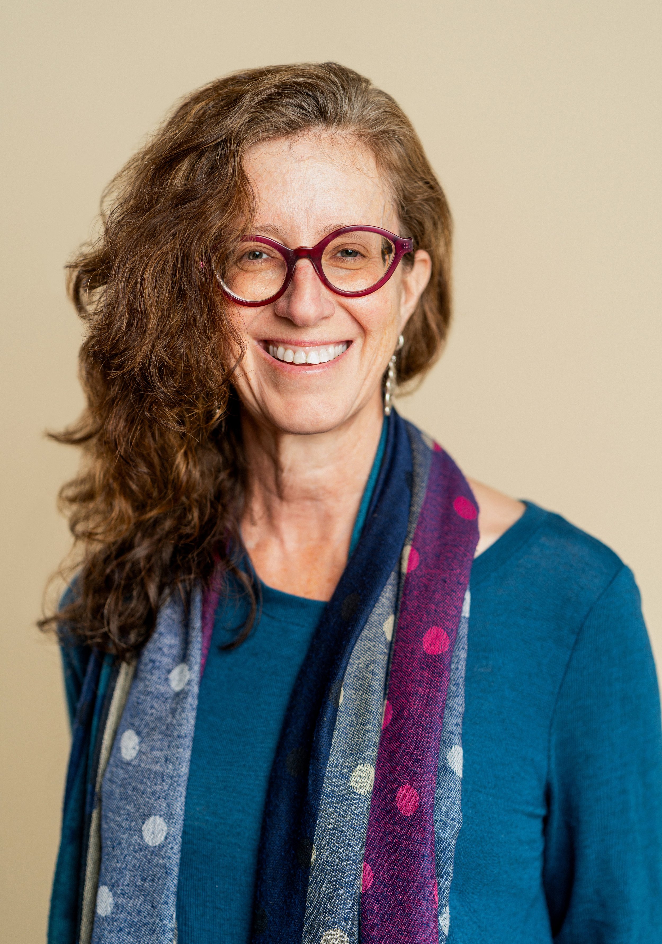 A woman with curly brown hair, wearing red glasses and a blue top, smiling with a colorful polka-dot scarf against a beige background.