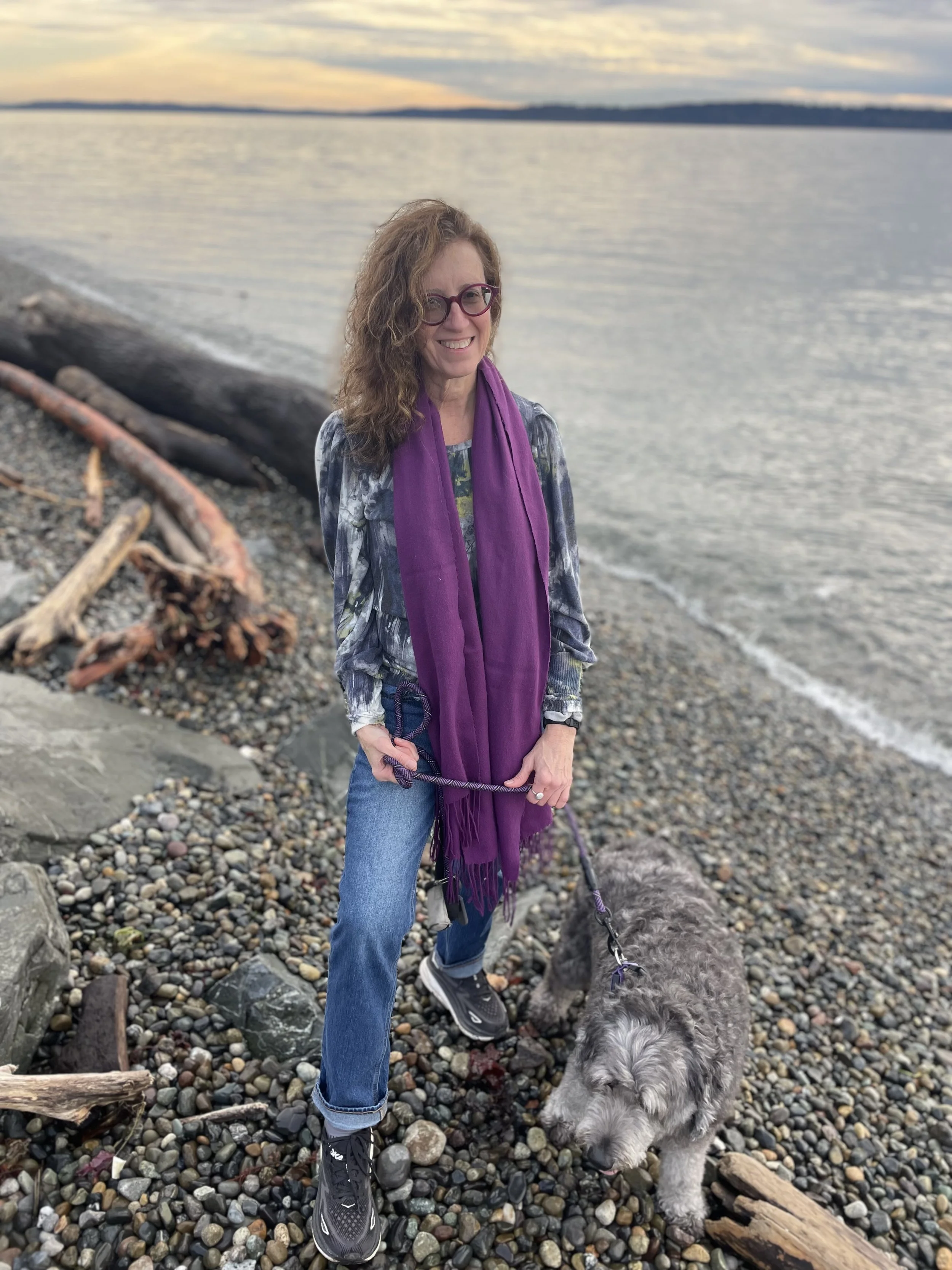 A woman with curly hair, wearing glasses, a patterned top, jeans, sneakers, and a purple scarf, standing on a rocky beach holding a leash attached to a shaggy gray dog, with a body of water and a sunset in the background.