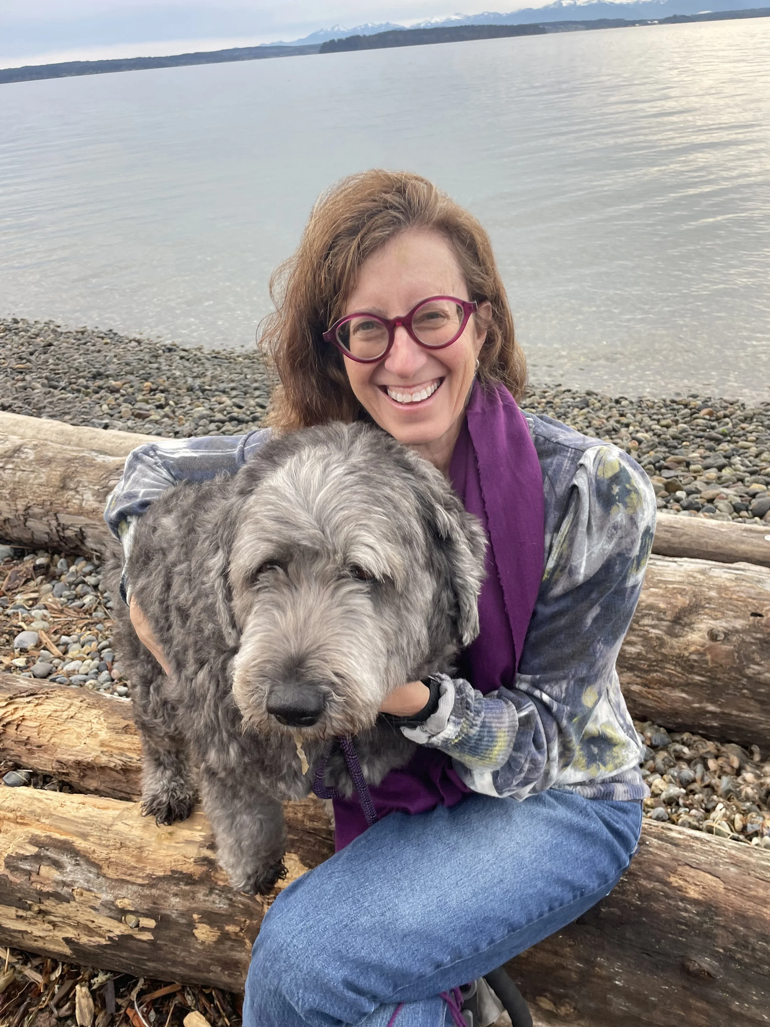 A smiling woman with curly hair and glasses sitting on a log by a rocky beach, holding an adorable gray and black fluffy dog with a wavy coat, with a body of water and mountains in the background.