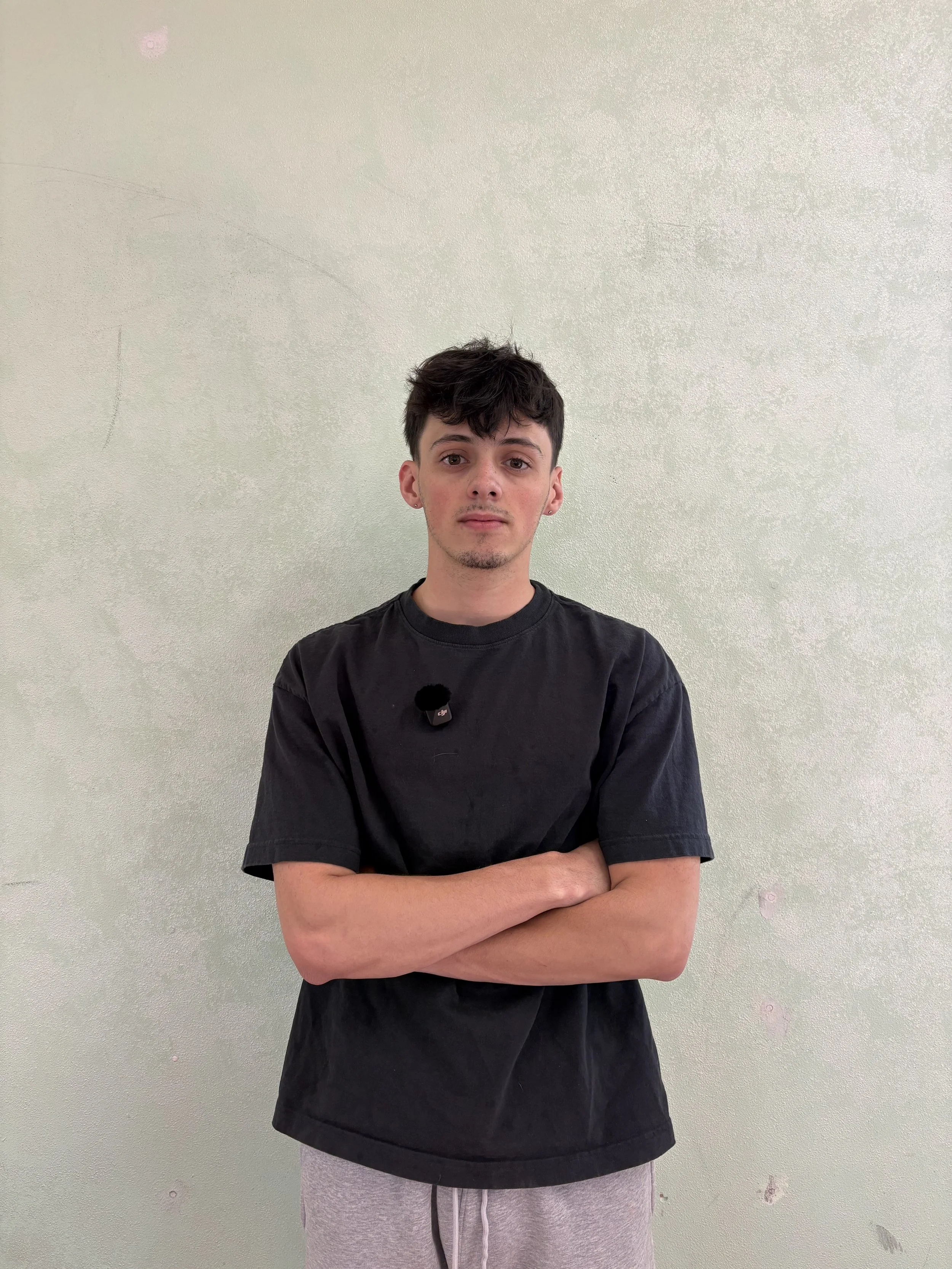 A young man with dark hair and light skin stands against a plain, light-colored wall with his arms crossed. He is wearing a black t-shirt and light gray sweatpants.