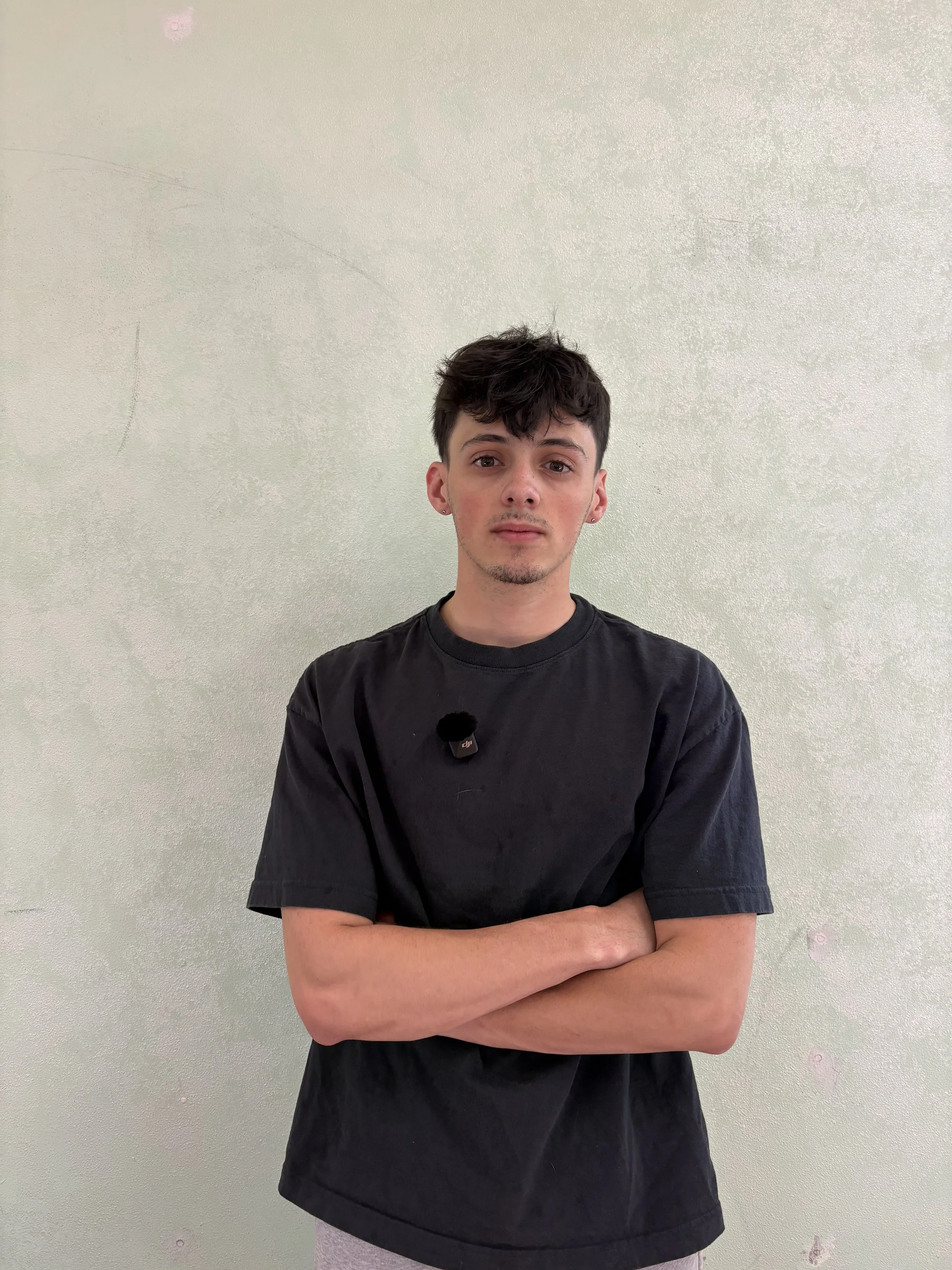 A young man with dark, tousled hair, wearing a black t-shirt, standing with his arms crossed in front of a light-colored wall.