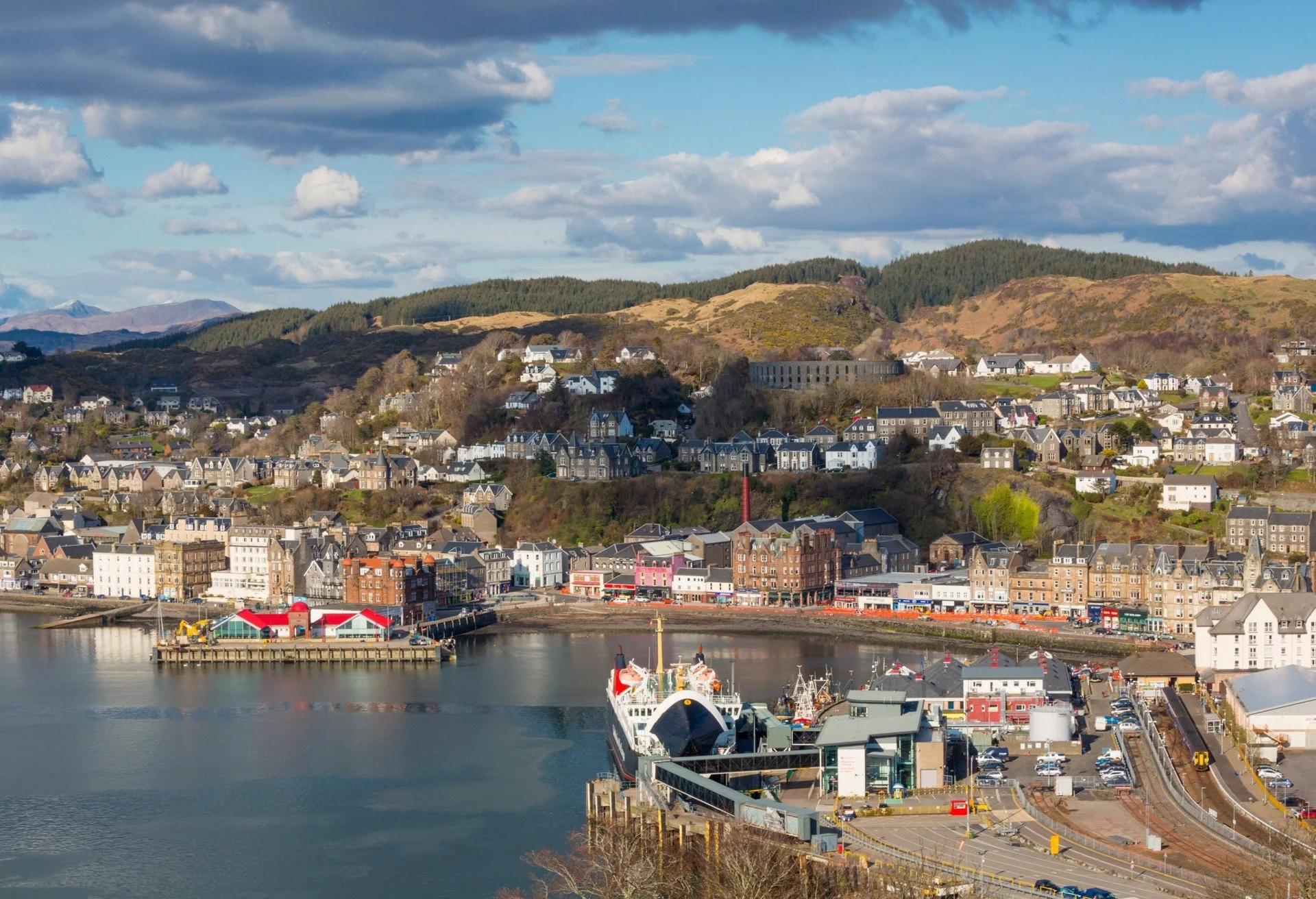 A coastal town with a harbor, docked ships, and colorful buildings at the waterfront. Houses and apartment buildings are on hills behind. Green hills and mountains are in the background under a partly cloudy sky.