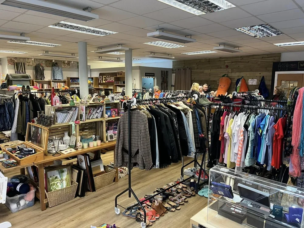 Inside a thrift store with clothing racks filled with jackets, shirts, and dresses, shelves with books, and various accessories and household items on display.