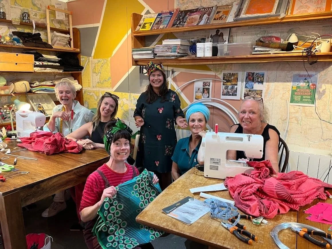 Group of seven women, some with headscarves, gathered around a sewing table in a craft room, smiling at the camera. There are sewing machines, fabric, and scissors on the table.