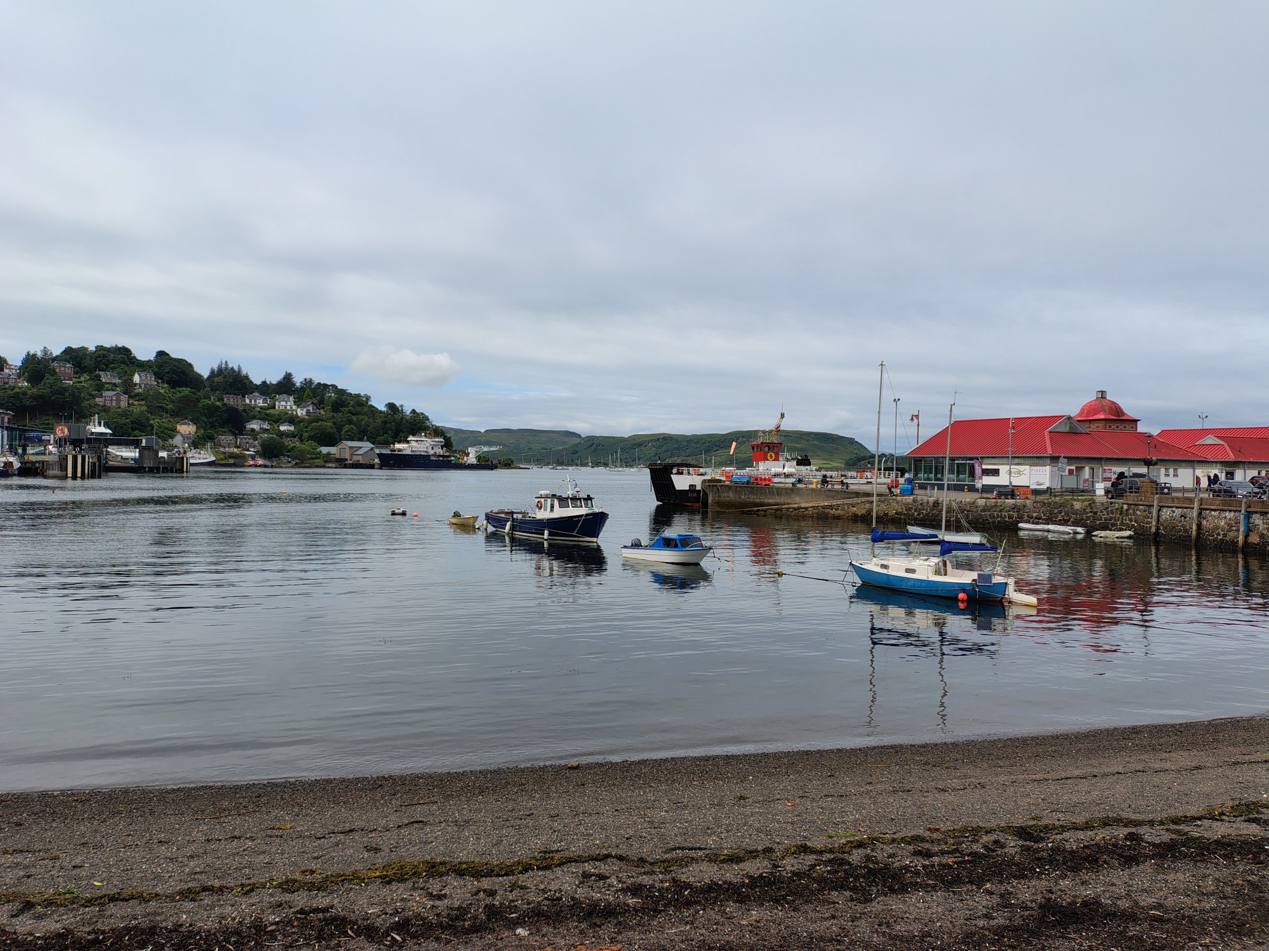 A harbor scene with boats docked along the pier, a hill with houses and trees in the background, and a building with a red roof and a small tower on the right side.