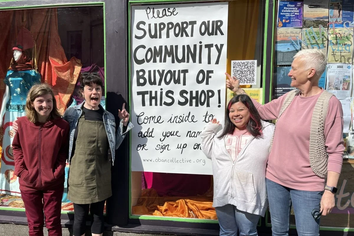 Four women standing together in front of a shop window with a large sign supporting the community. One woman, with short gray hair and wearing a pink sweater and a knitted vest, is pointing at the sign. The other three women are smiling and gesturing, with one wearing a denim jacket, one in a white hoodie, and one in a maroon outfit.