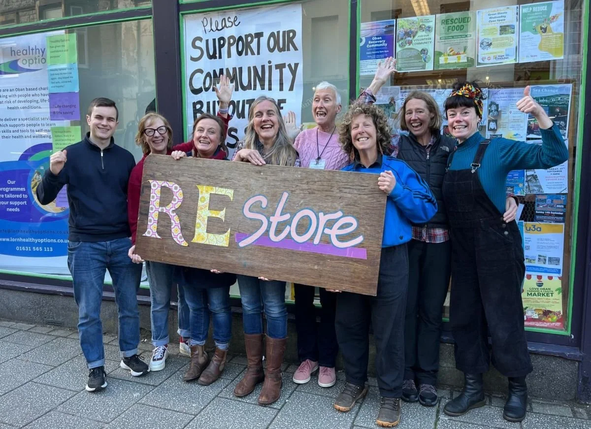 Group of nine people standing on a sidewalk in front of a building with windows, holding a large wooden sign that says 'REstore' in colorful letters, smiling and waving.
