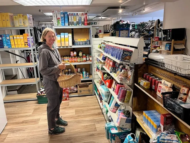 A woman with blonde hair holding a basket in a grocery store aisle, surrounded by shelves stocked with food and household items.