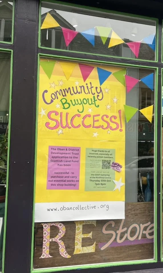 Sign on storefront celebrating community success with colorful bunting and posters. The sign reads "Community Buoyed SUCCESS!" and includes information about local development trust and a community gathering event.