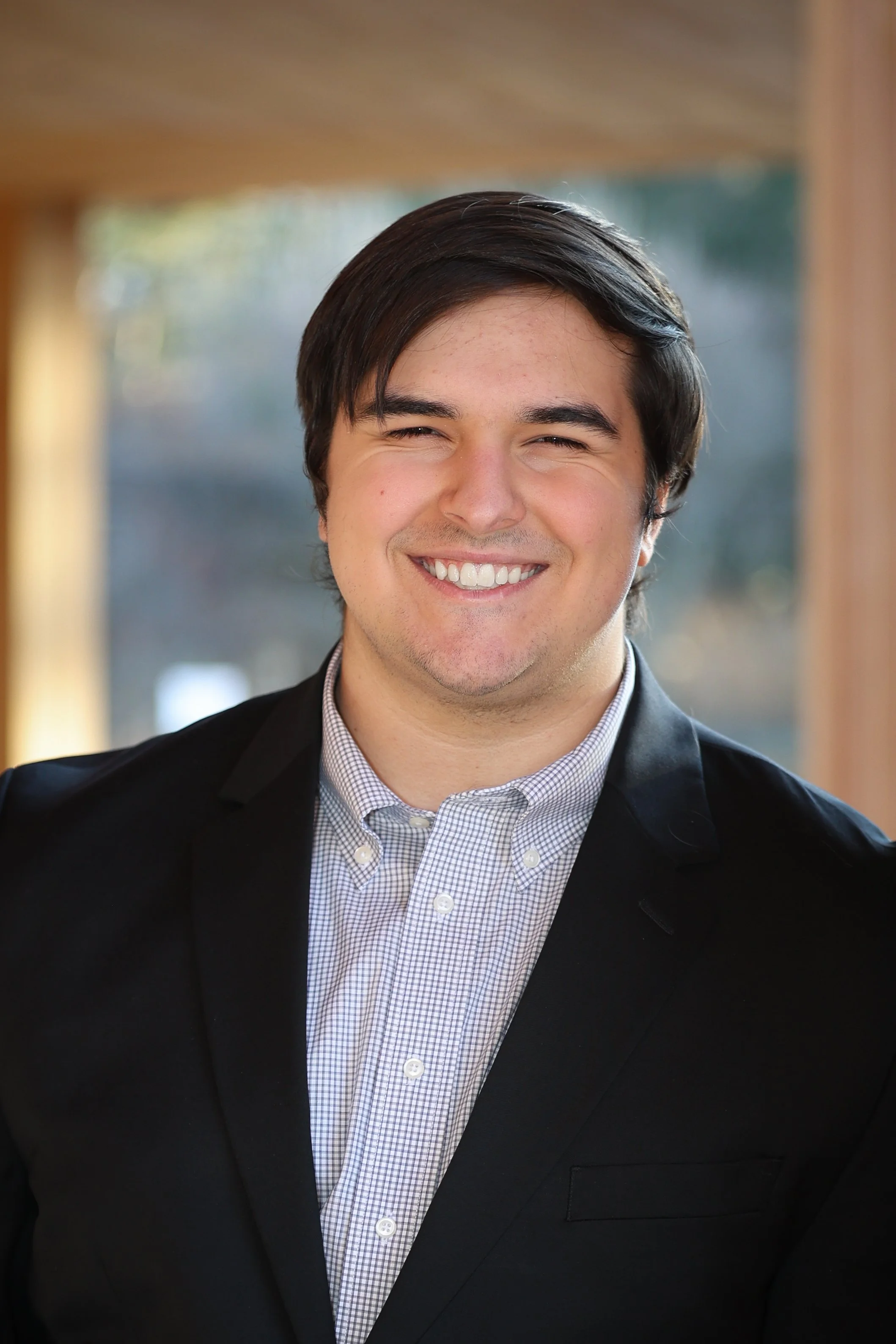 A young man with dark hair and a big smile, dressed in a checkered shirt and dark blazer, standing indoors in front of blurred background with wooden and glass elements.