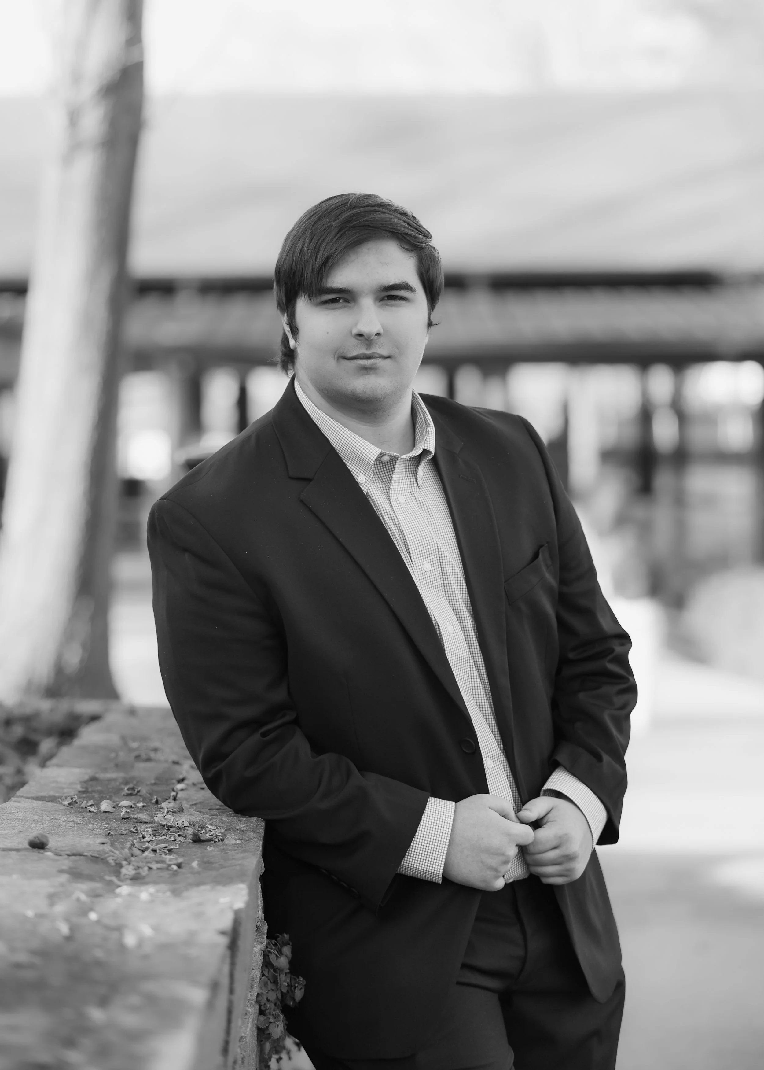 A young man in a suit leaning on a low wall outdoors, with a bridge and river in the background.