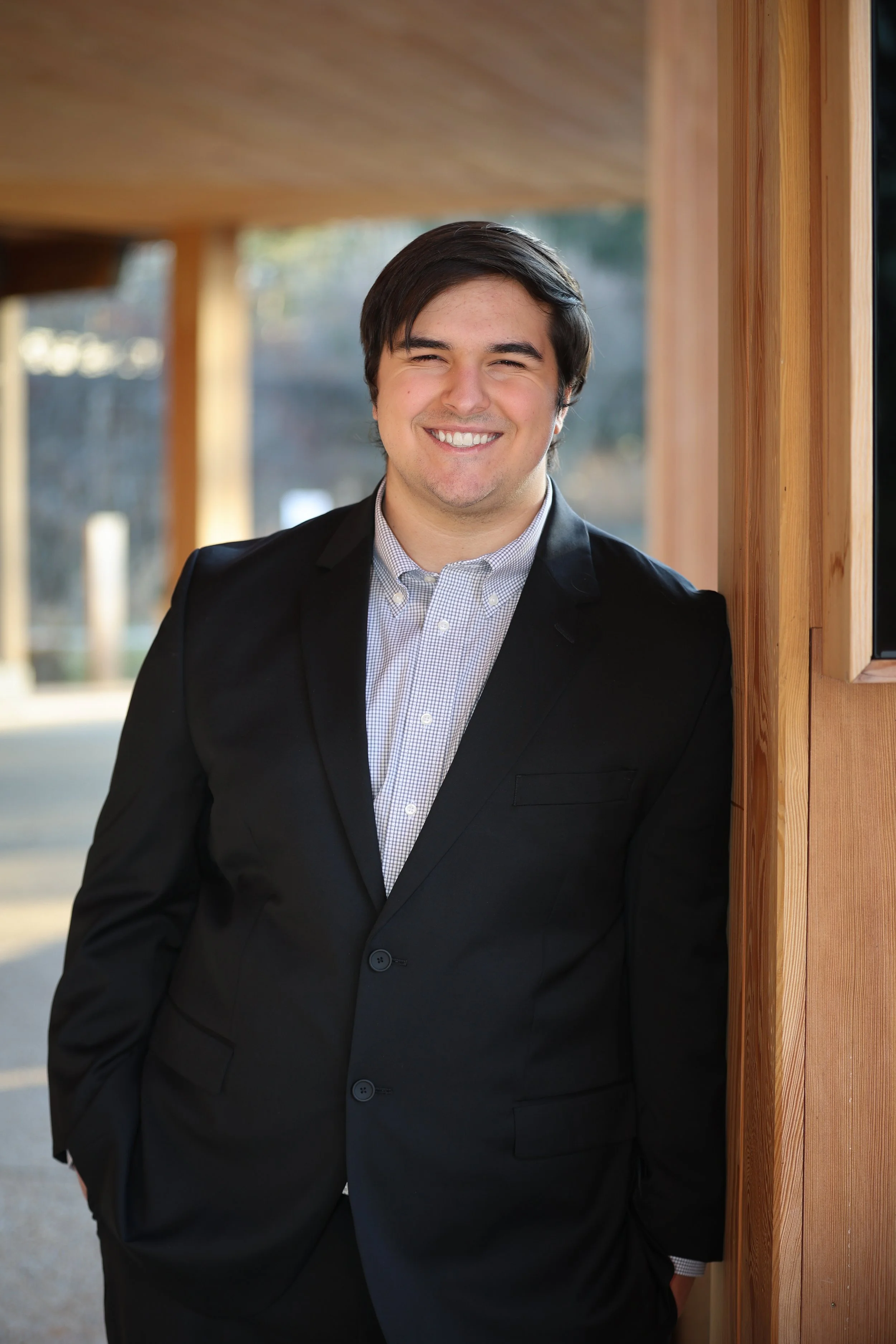 A young man in a black suit and light checkered shirt, smiling, standing outdoors next to a wooden wall.
