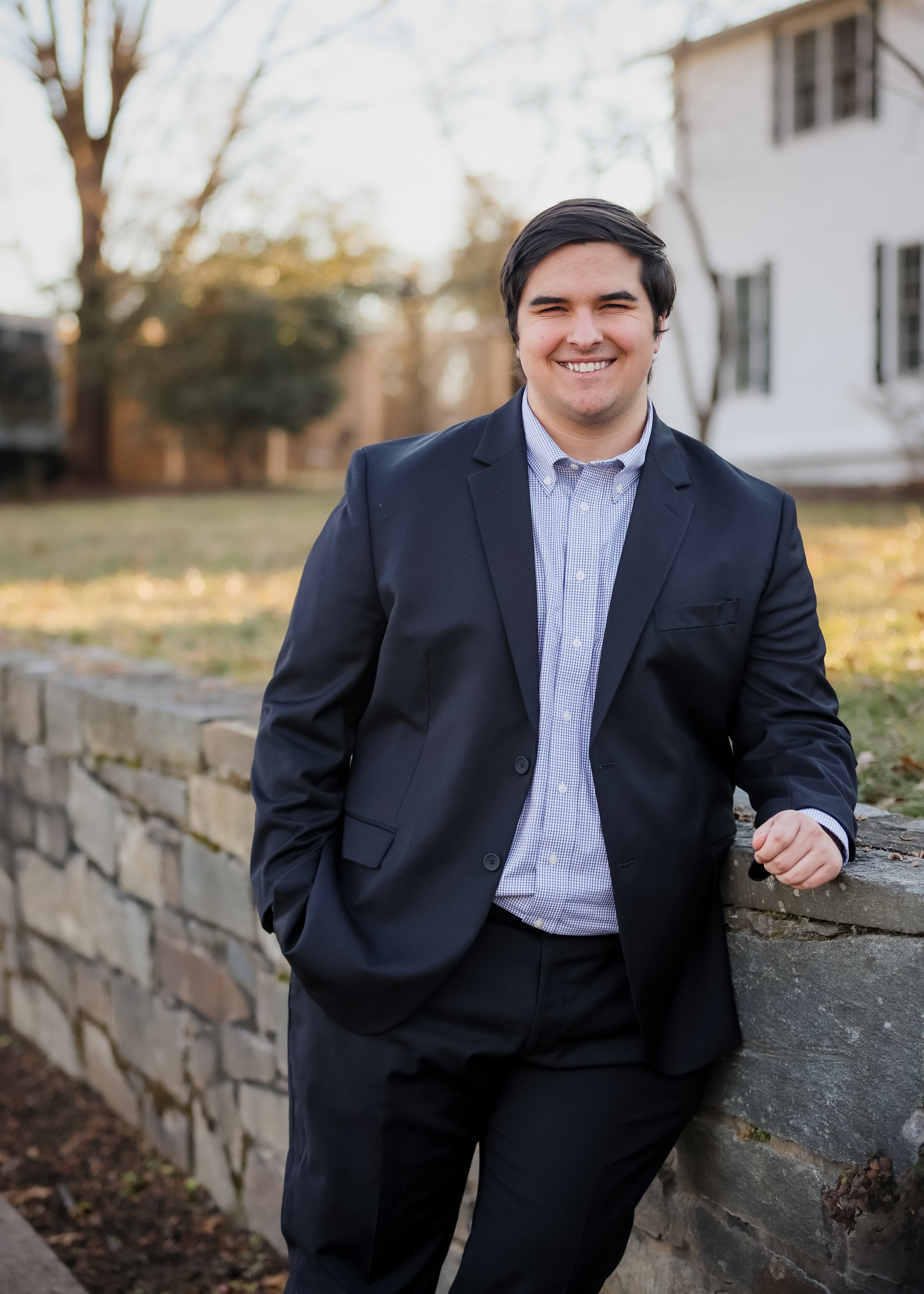A young man in a navy blazer and blue dress shirt leaning on a stone ledge outdoors, smiling, with trees and a white building in the background.