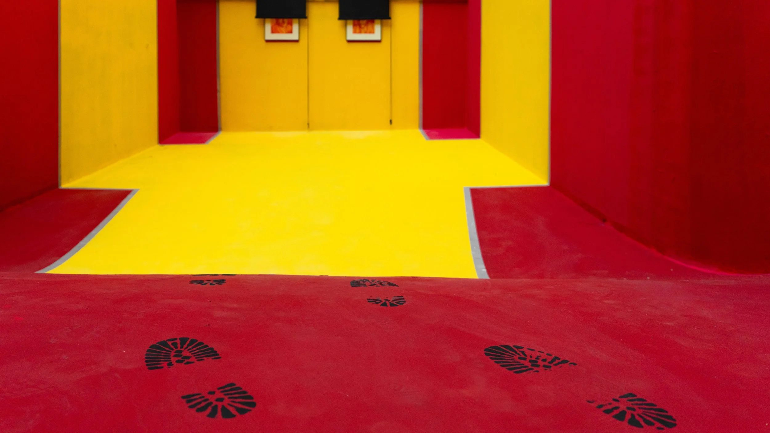 Colorful indoor slide with red sides, yellow sliding surface, and black footprint decals on the red area at the top, leading down into a red and yellow room with framed pictures on the wall.