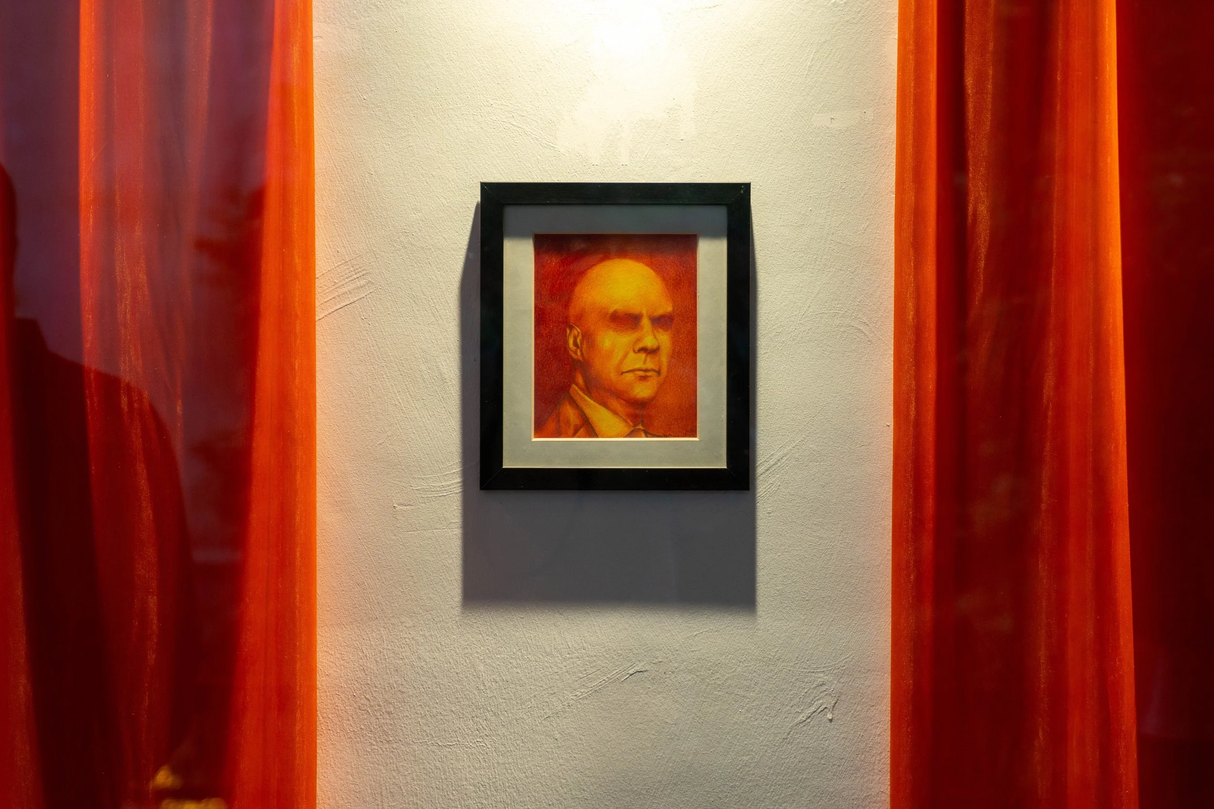 Framed painting of a man with a bald head, wearing a suit and tie, hanging on a beige wall between red curtains.