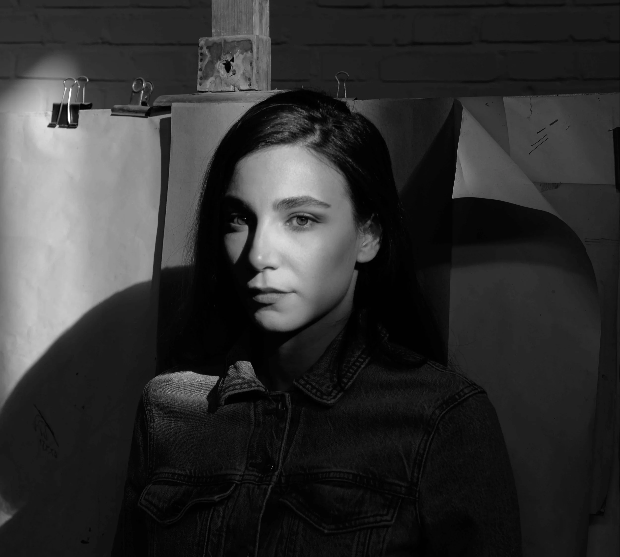 Black and white photo of a young woman with dark hair in a denim jacket, standing in front of a wall with paper and clipboards, casting a shadow on her face.