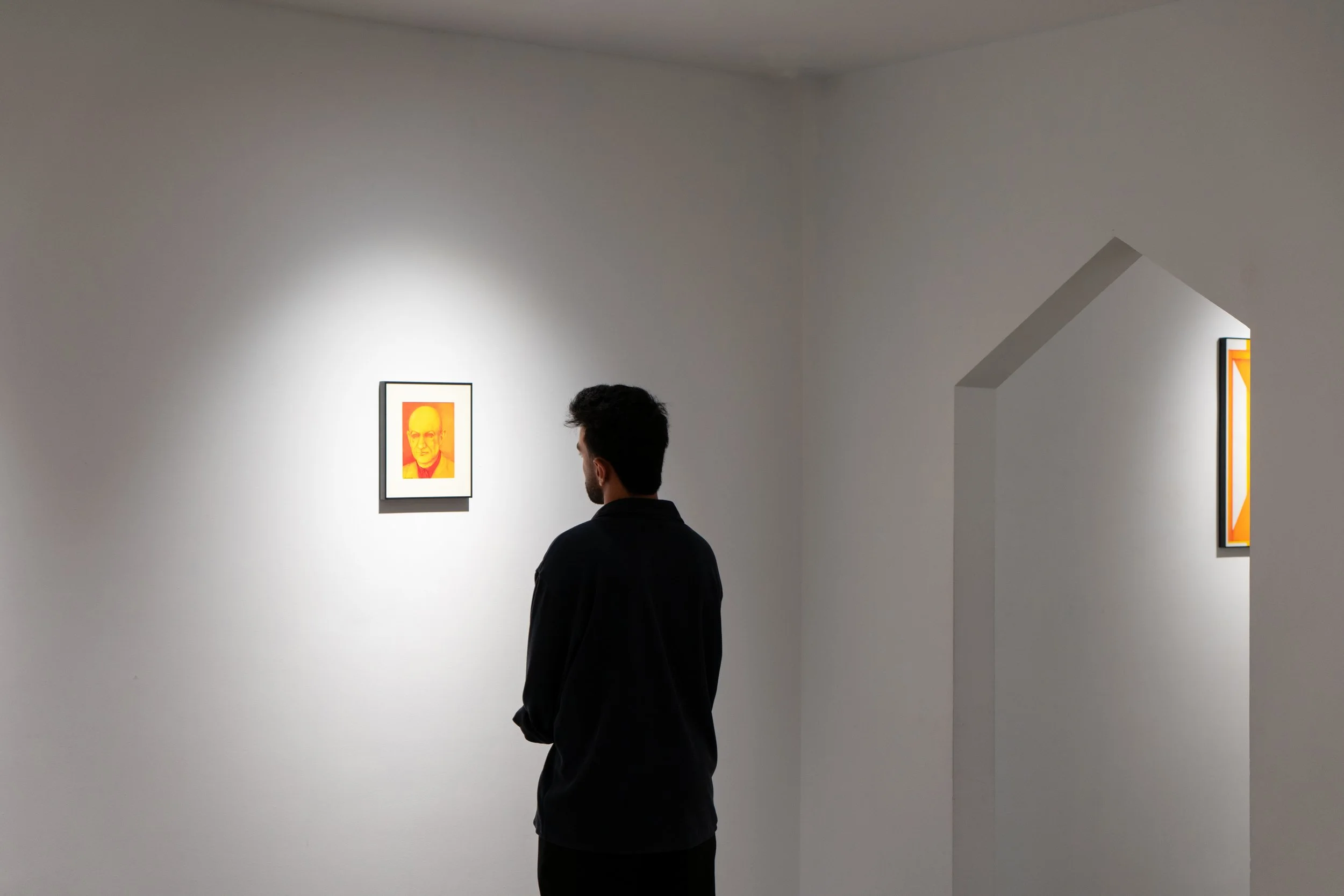 A man with dark hair and a beard, wearing a black shirt, stands in an art gallery viewing a framed portrait on a white wall. The portrait features a person's face rendered in yellow and orange hues.