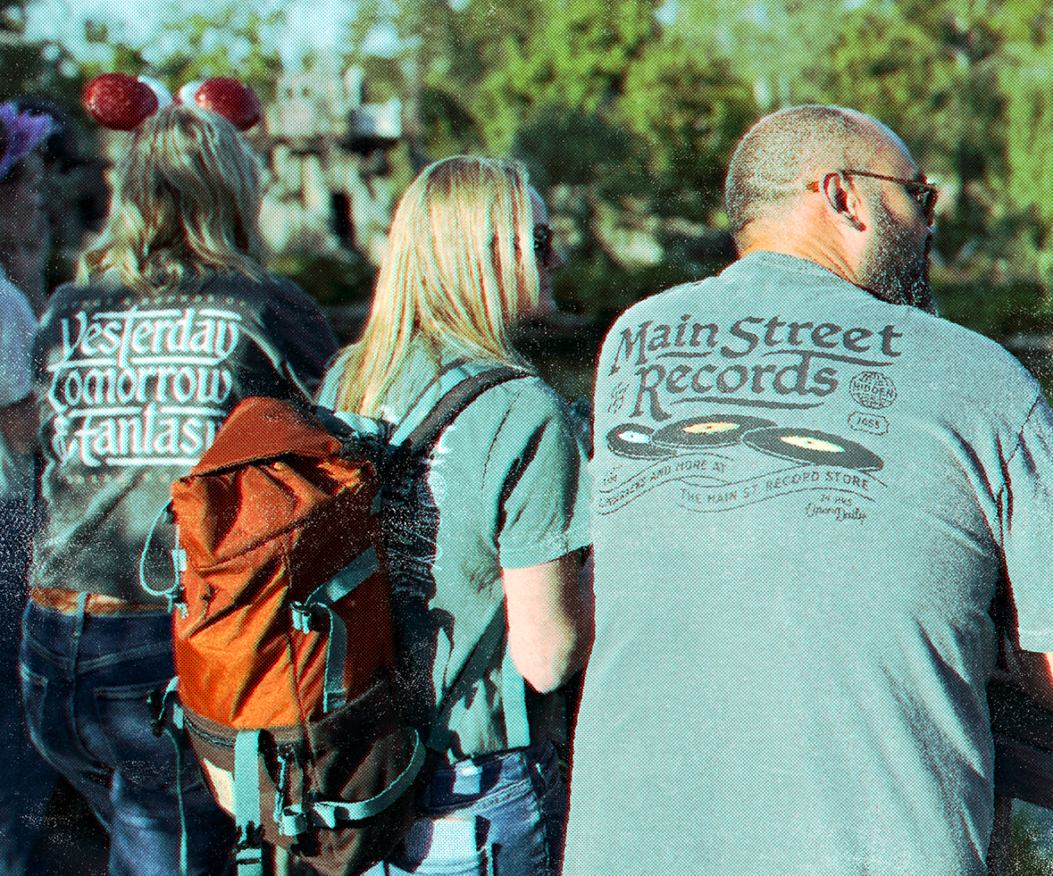 Three people standing outdoors, with one woman wearing a brown backpack, and two men, one with glasses and a tattoo on his arm, the other with headphones around his neck. It appears to be a festival or outdoor event.