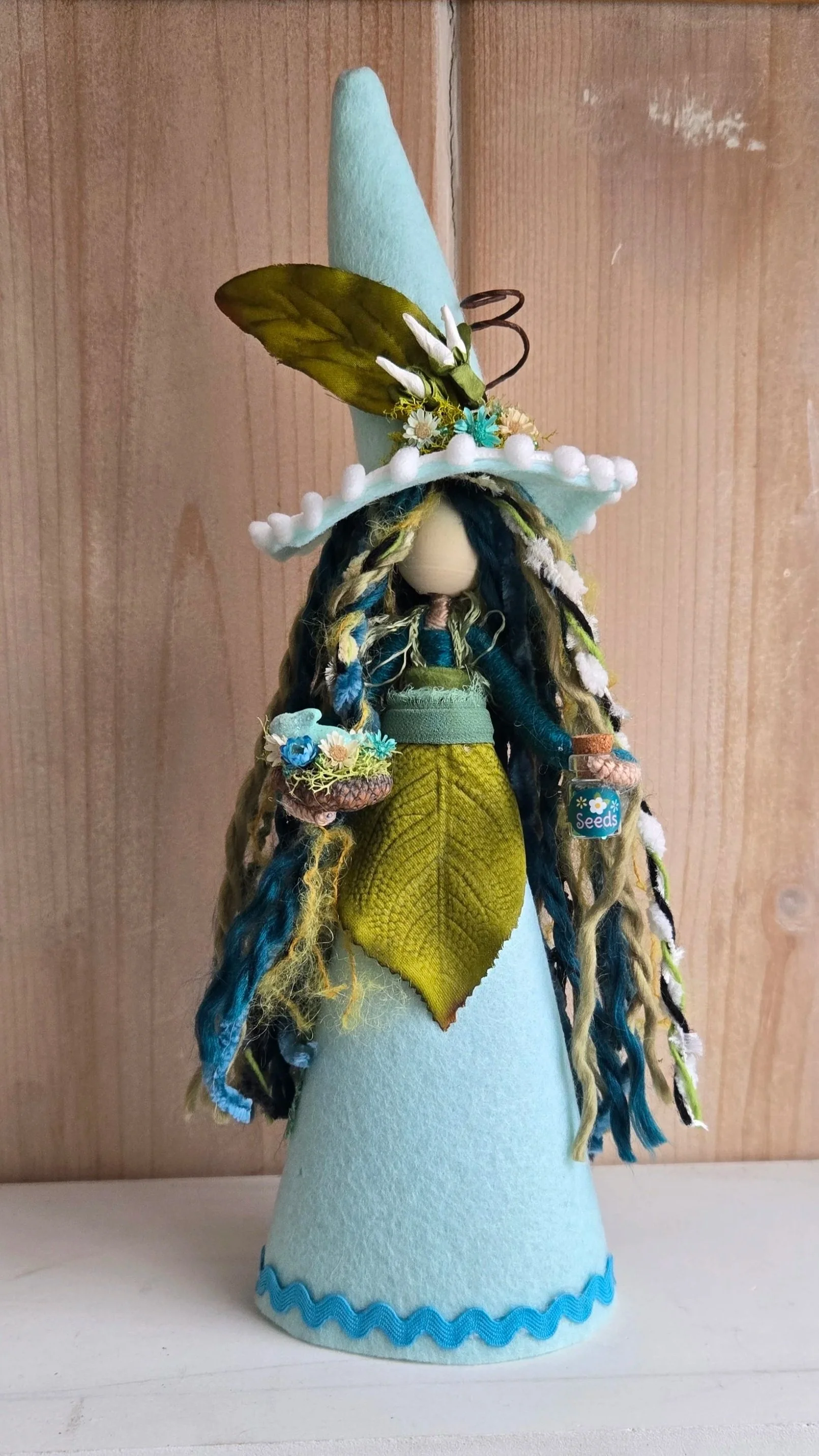 Handmade doll dressed as a witch with long yarn hair and a tall blue felt hat with spring floral decoration, standing on a white surface against a wooden background.