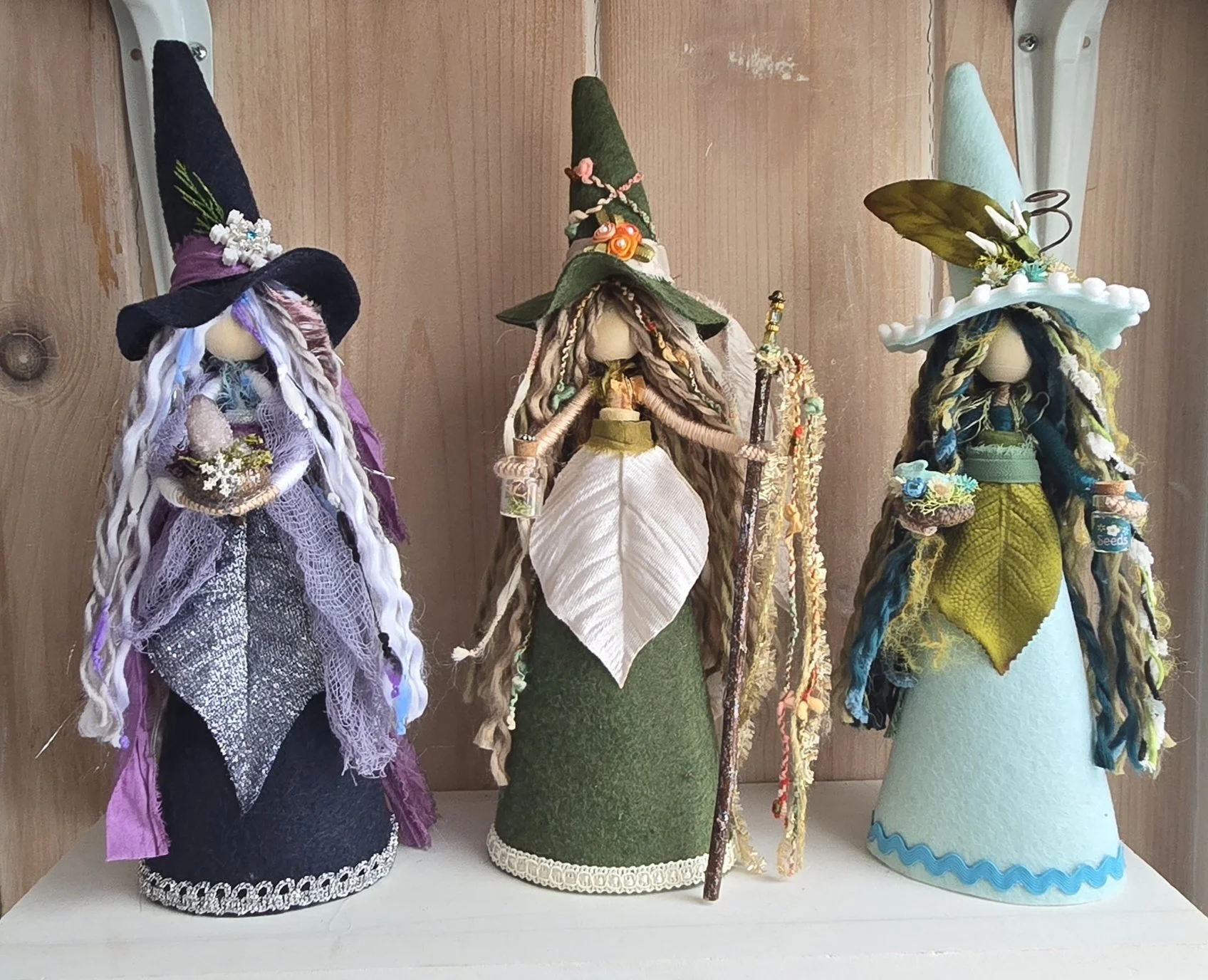 Three handcrafted witch dolls with elaborate costumes and accessories, each wearing a pointed hat, displayed on a white shelf against a wooden background.