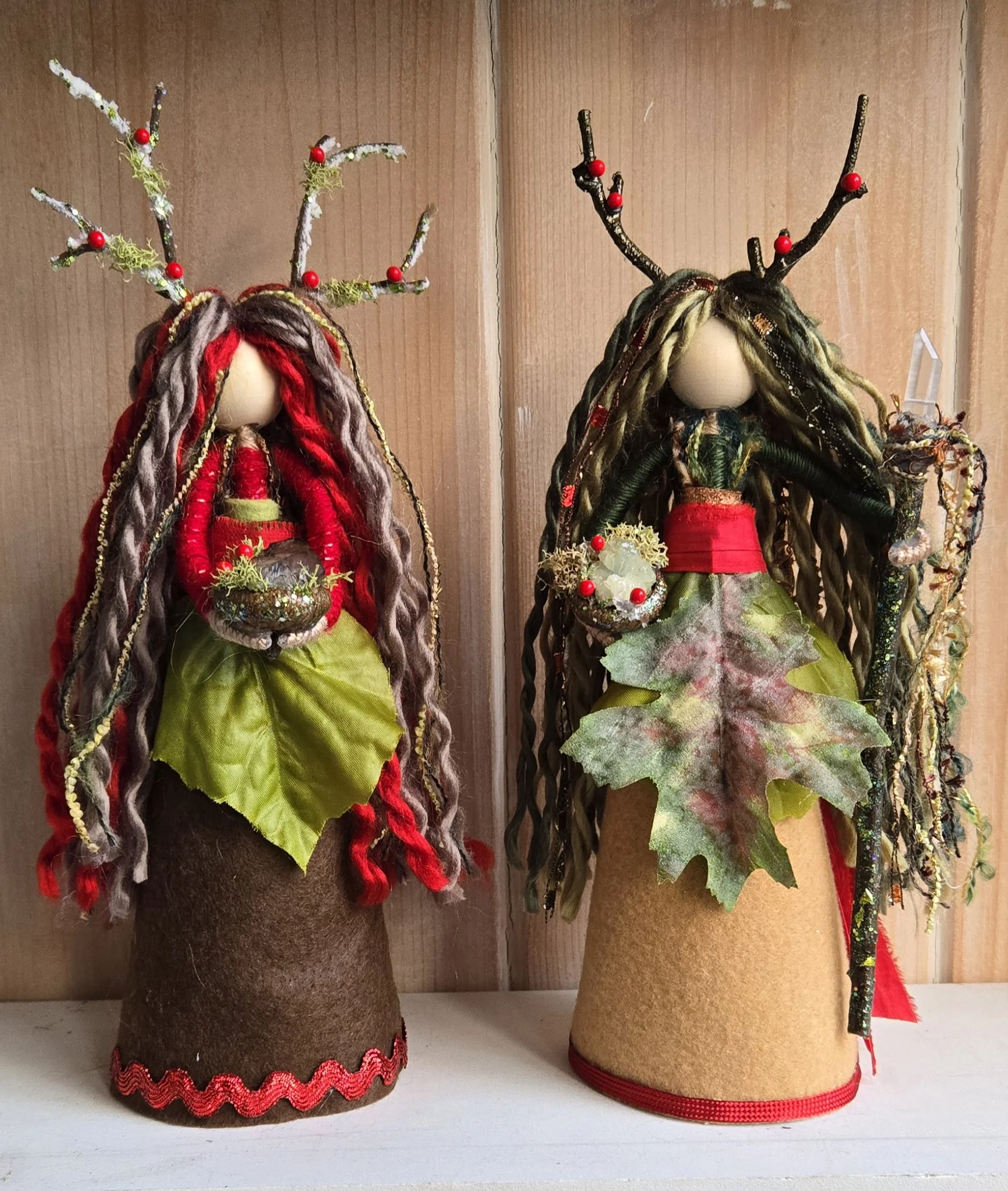 Two handcrafted Christmas dolls with antlers, dressed in festive colors, with felted bases, placed on a white surface against a wooden background.