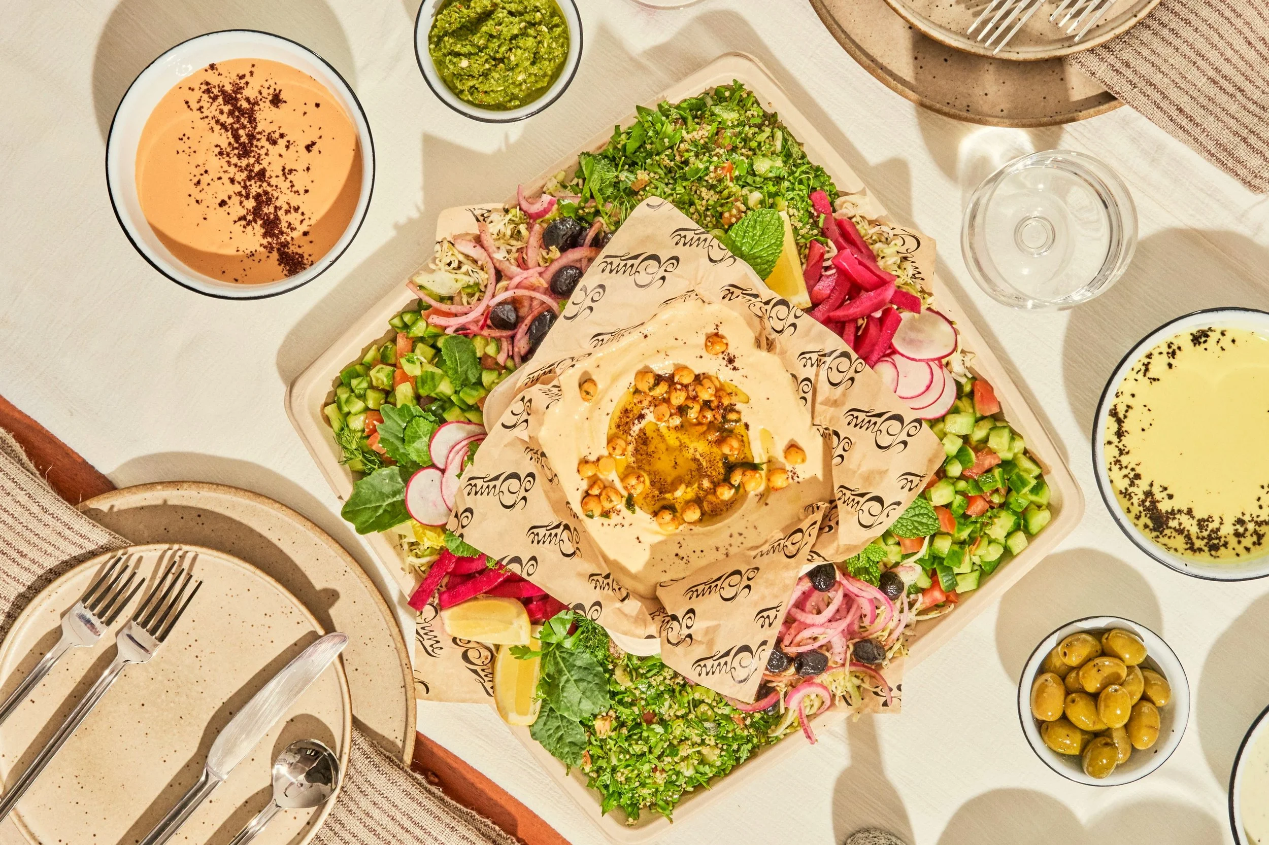A Middle Eastern mezze platter with hummus topped with chickpeas and olive oil, surrounded by fresh vegetables, salads, pickles, and sauces, along with glasses of white wine or champagne, on a white table.