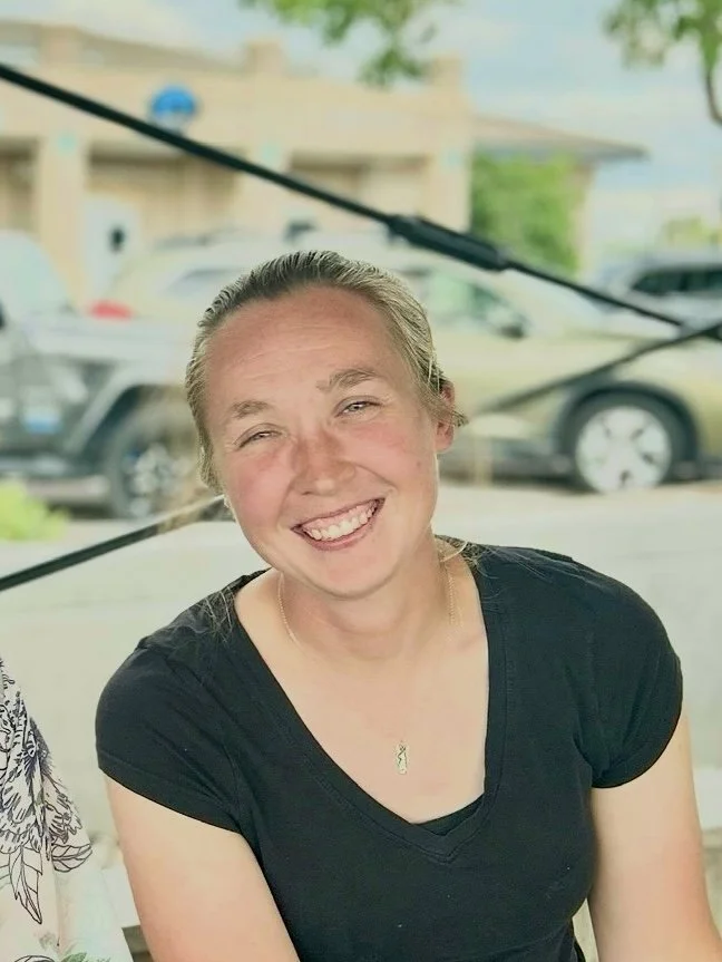 A smiling woman with blonde hair tied back, wearing a black shirt and a gold necklace, sitting outdoors at a restaurant or café with a blurred background of cars and trees.