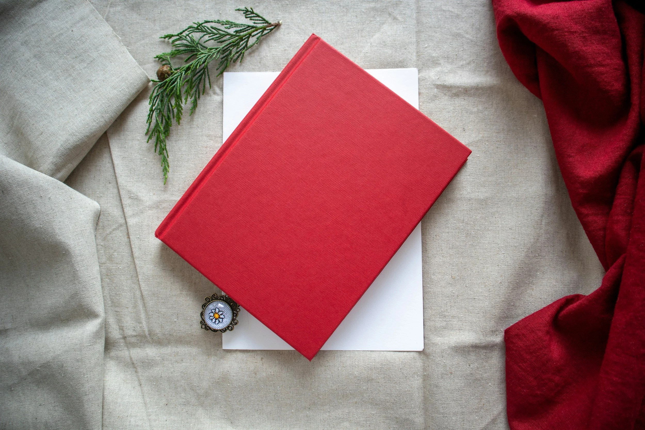 Red hardcover book on white paper, surrounded by beige and red cloths, with a sprig of green pine and a small decorative item with a flower design nearby.