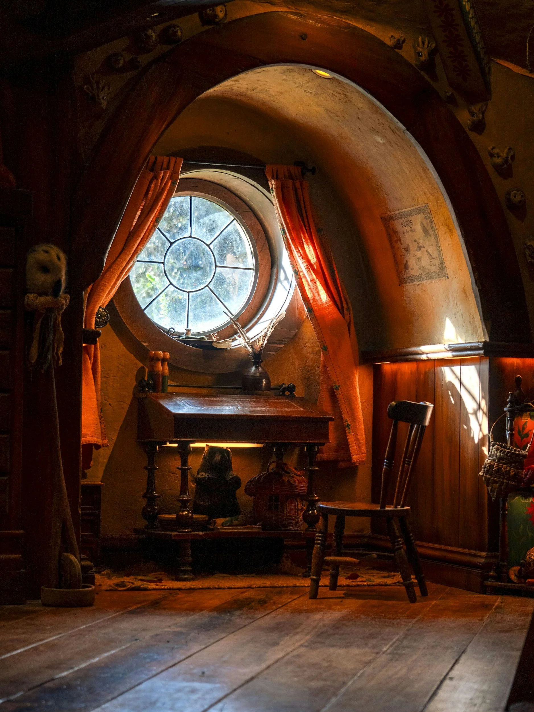 Cozy room with round window, orange curtains, wooden furniture, and decorative items, illuminated by natural light.