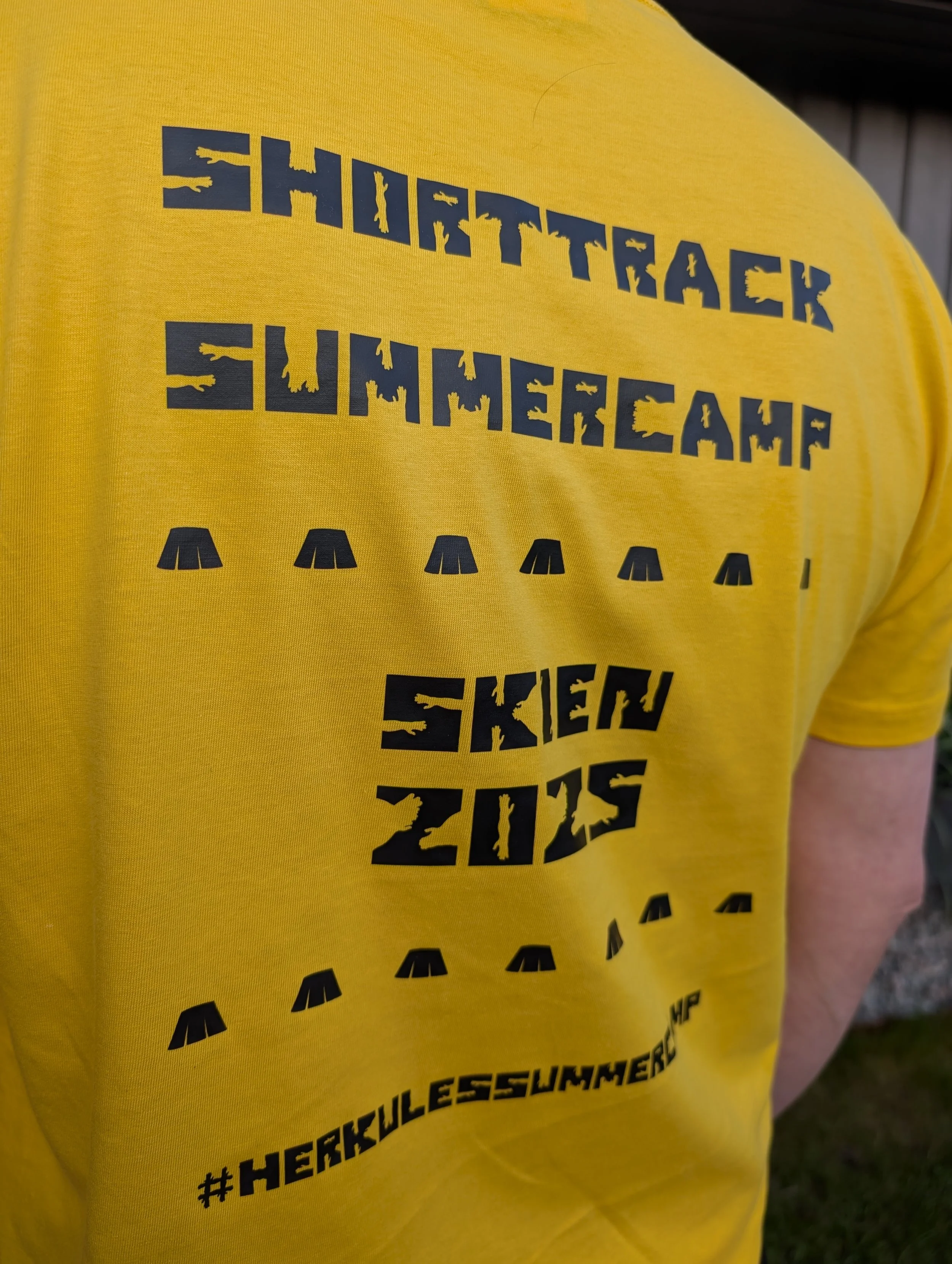 Back of a yellow t-shirt with black text that reads 'SHIRTTRACK SUMMER CAMP 2019 SKIEW' and hashtags '#HERKULLESSUMMER'.